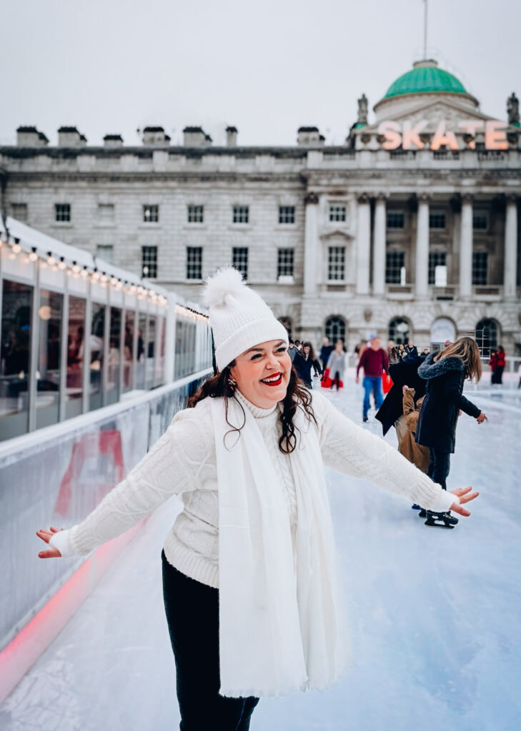 Brand photographer Rosie Parsons ice skating at Somerset House in London wearing a cozy christmas outfit of a white cable knit sweater, cream pom pom beanie, white scarf and black trousers, smiling with arms outstretched on the ice rink