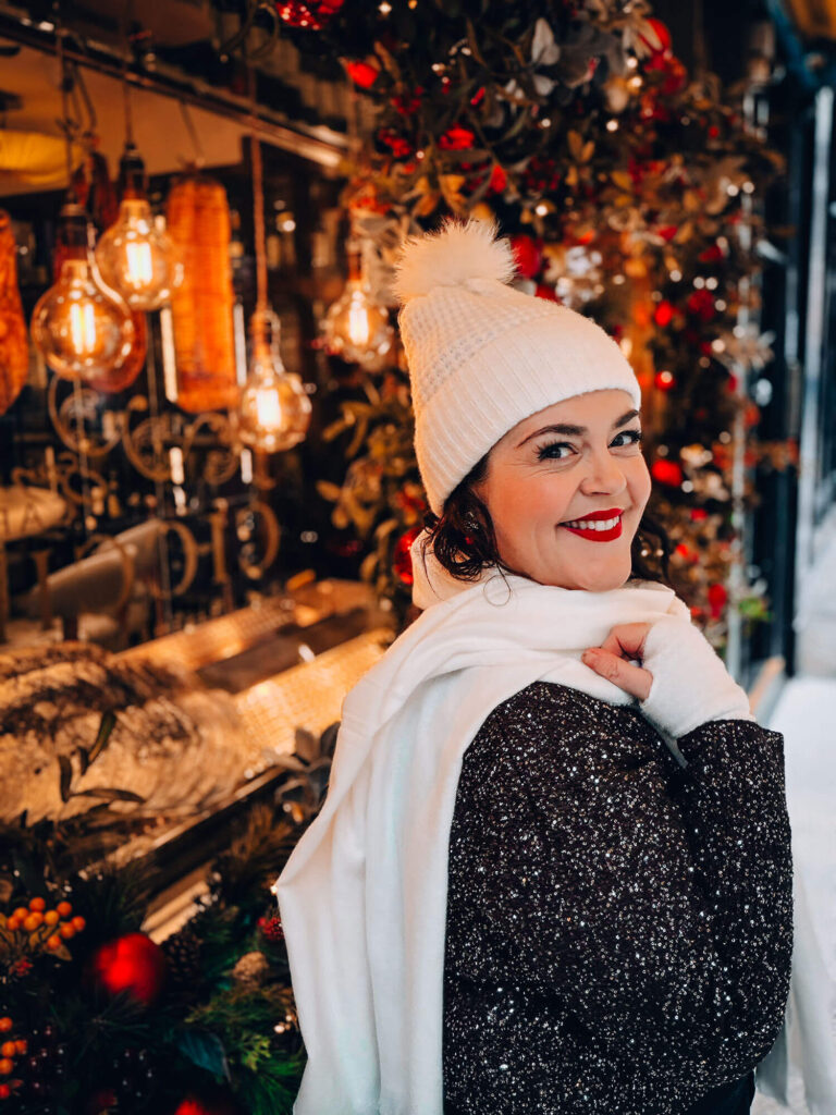 Business blogger Rosie Parsons next to a festive Christmas window wearing a sparkly black jumper, cream knit beanie with pom pom, and white scarf, surrounded by warm festive lights and red decorations