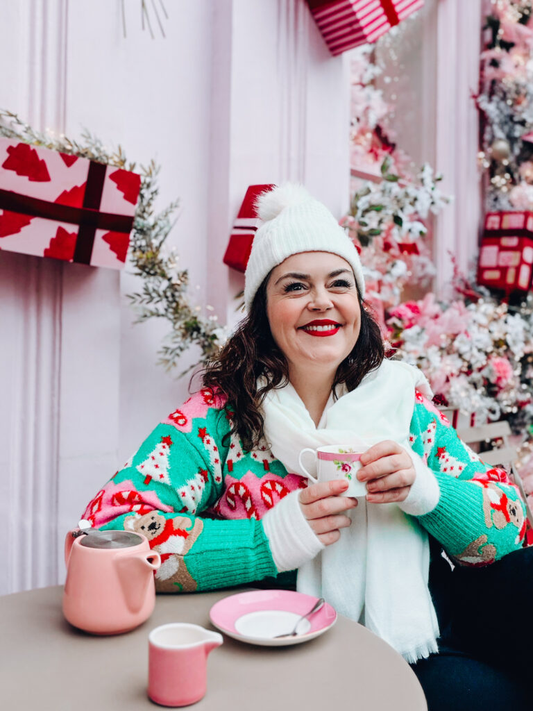Professional photographer Rosie Parsons enjoying a casual christmas party outfit moment in a pink themed cafe, wearing a colourful festive jumper with teddy bears and candy canes, white pom pom beanie and cream scarf, holding a cup of tea surrounded by pink Christmas trees and wrapped gifts