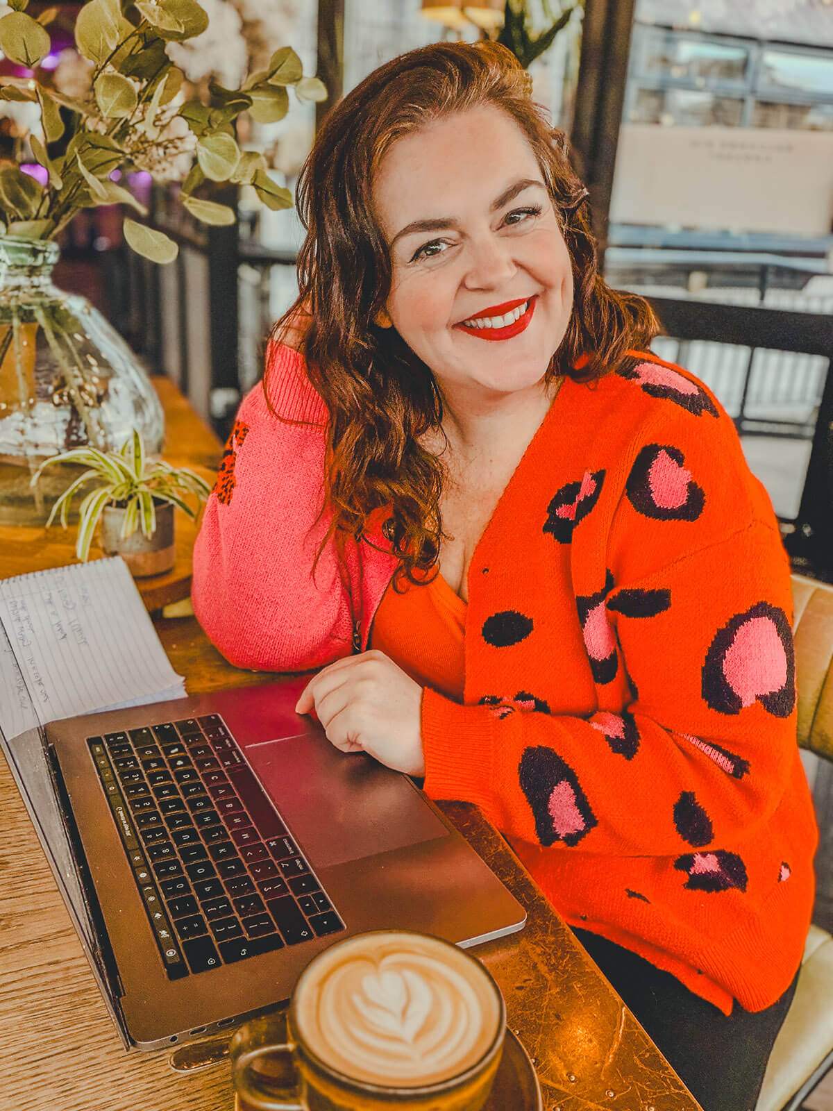 Rosie Parsons, a small business blogger, smiles warmly while sitting at a wooden table in a cosy coffee shop. She wears a bright orange cardigan with pink and black leopard print and has auburn wavy hair and red lipstick. In front of her is an open laptop and a latte with heart foam art. Green eucalyptus stems in a vase add a fresh touch to the background. She's researching the best online course website platforms and course website design options for creators.