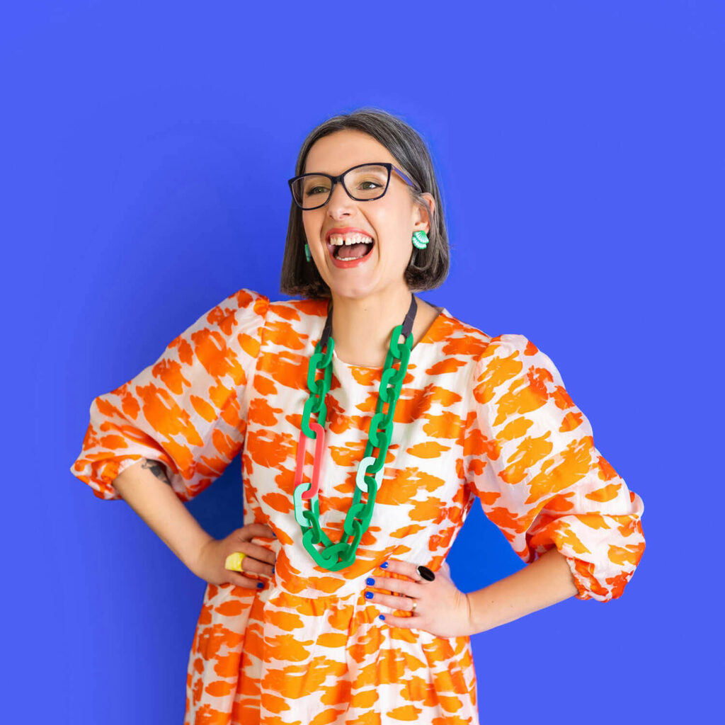 Confident woman laughing during a colourful business photoshoot, wearing a bold patterned dress and statement accessories against a bright backdrop.