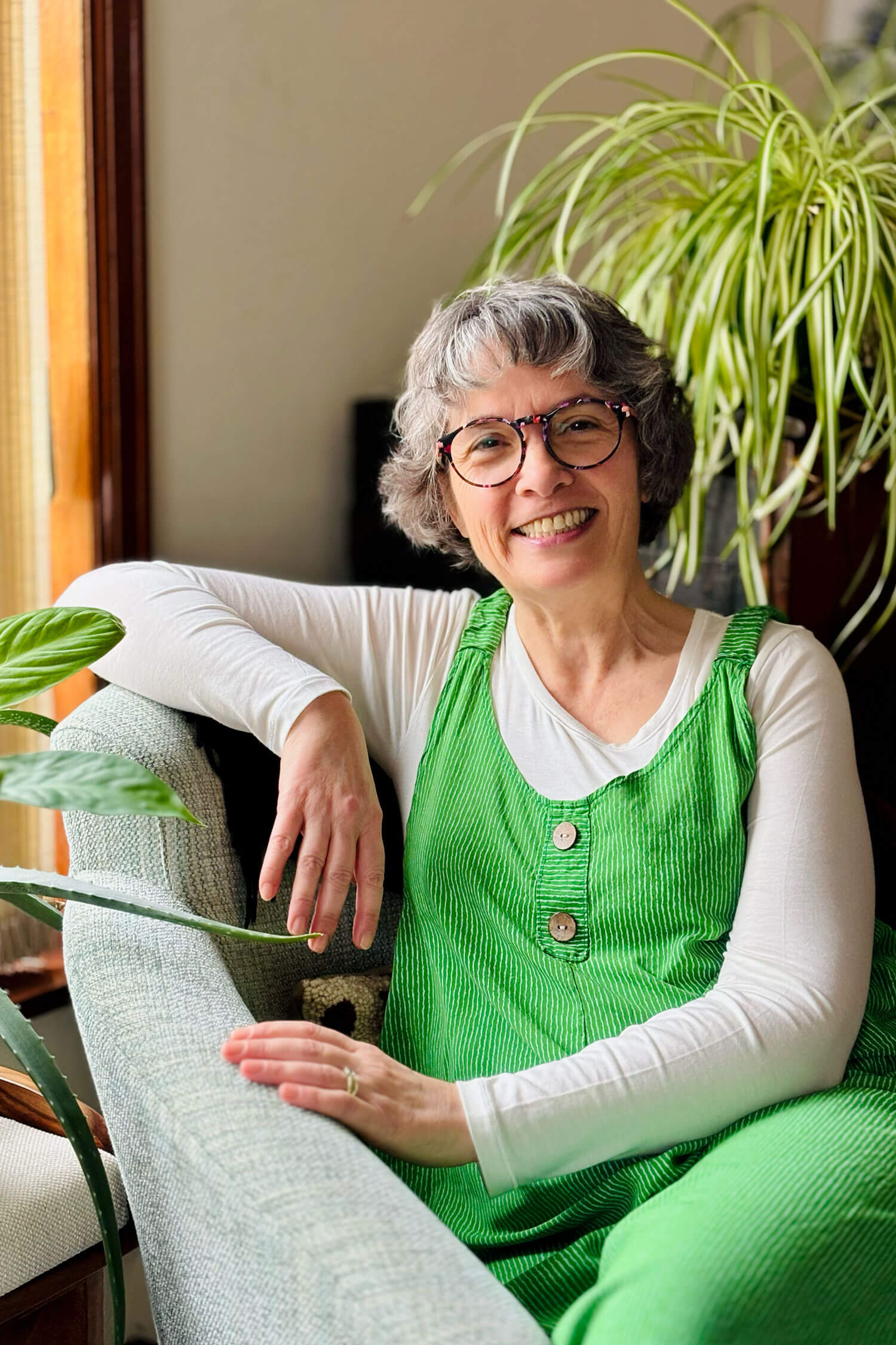 A female therapist sits comfortably on a sofa near houseplants, smiling warmly in a green pinafore and glasses, photographed for a relaxed self portrait business photoshoot.