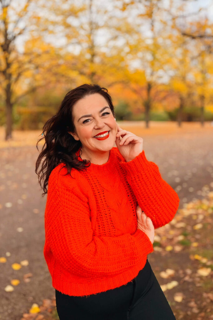 A woman in a bright orange jumper smiles warmly in a park filled with golden autumn leaves, captured in a relaxed outdoor portrait.