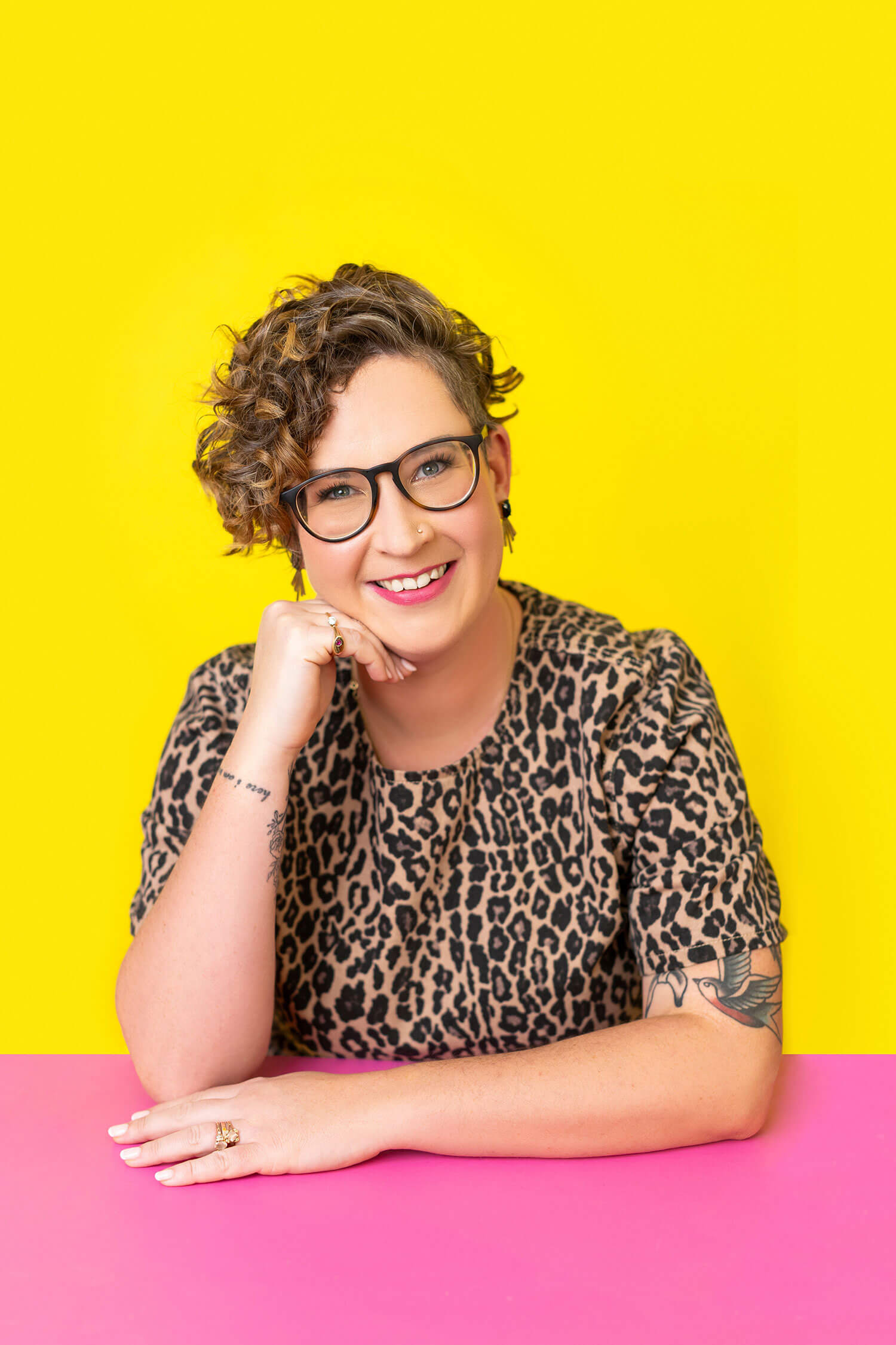 A woman with short curly hair and glasses smiles with her chin resting on her hand, sitting at a pink table against a bold yellow background.
