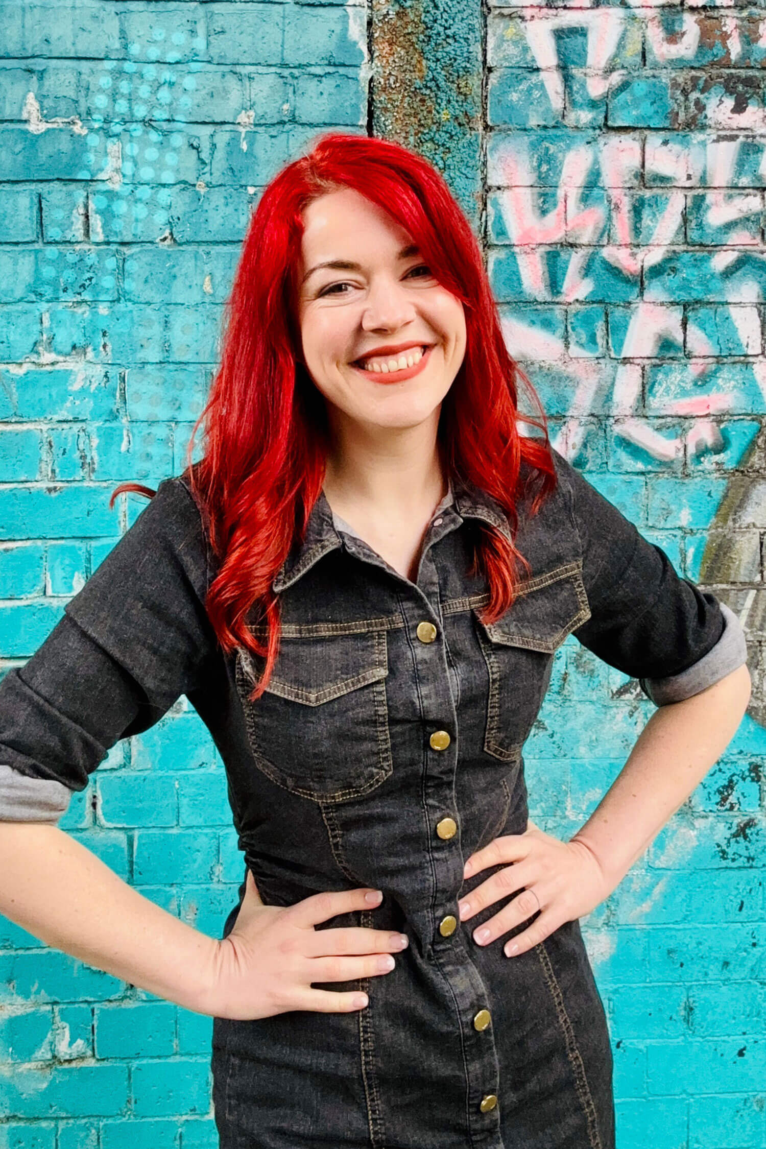 A woman - Heather Murray from AI for Non Techies - with bright red hair smiles confidently with her hands on her hips, standing in front of a turquoise graffiti wall in an urban setting. 