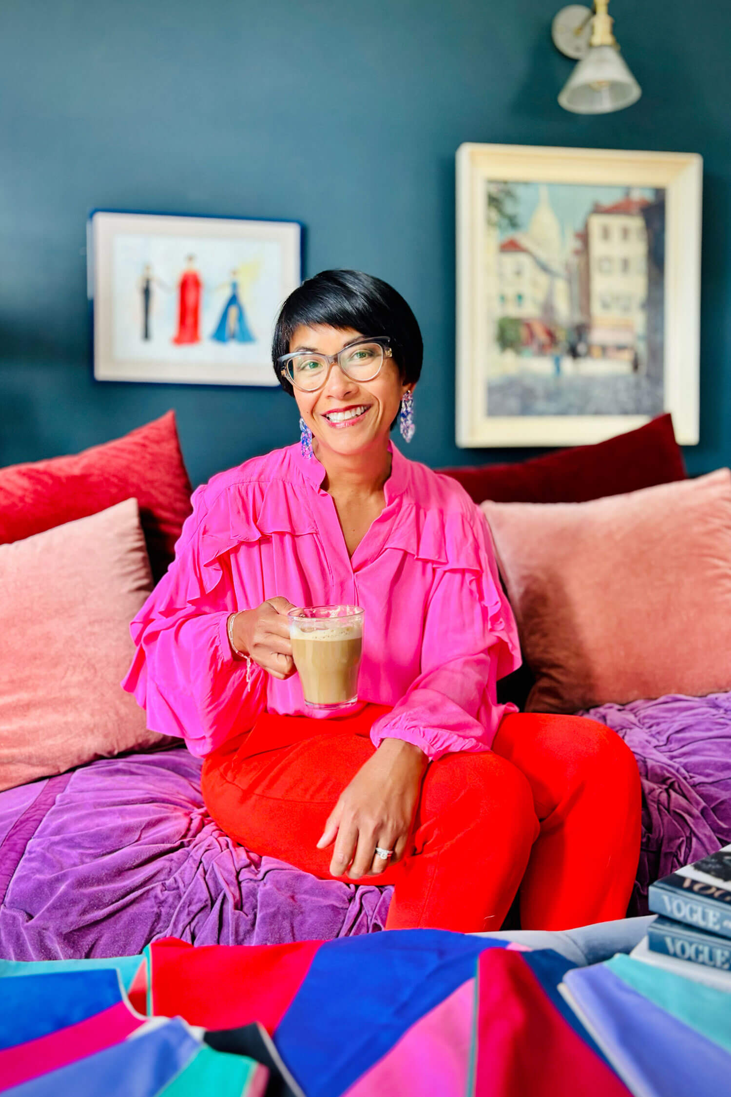 Personal stylist Chantelle Znideric in a bright pink blouse and red trousers sits on a colourful sofa holding a coffee, surrounded by fabric swatches and fashion books, smiling warmly in a relaxed setting.