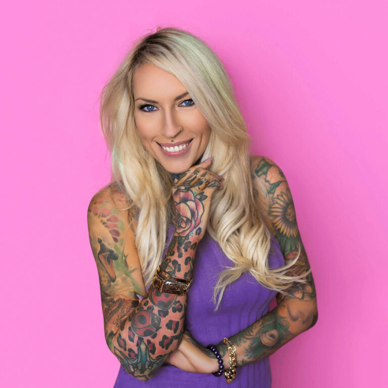 A woman - Lea Turner - with long blonde hair and colourful tattoo sleeves poses confidently against a bright pink background, smiling with one hand near her face. The clean, bold setup highlights expression, posture and styling ideas for professional business headshots.
