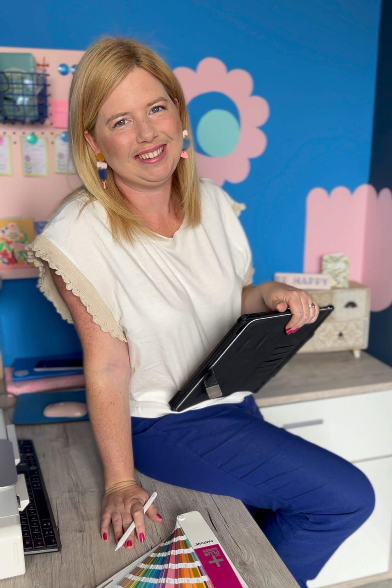 A woman - Kayleigh Lloyd from Lloyd Creative - sitting on a desk in a colourful studio, holding a tablet and smiling, with design tools and swatches beside her during a business photoshoot.