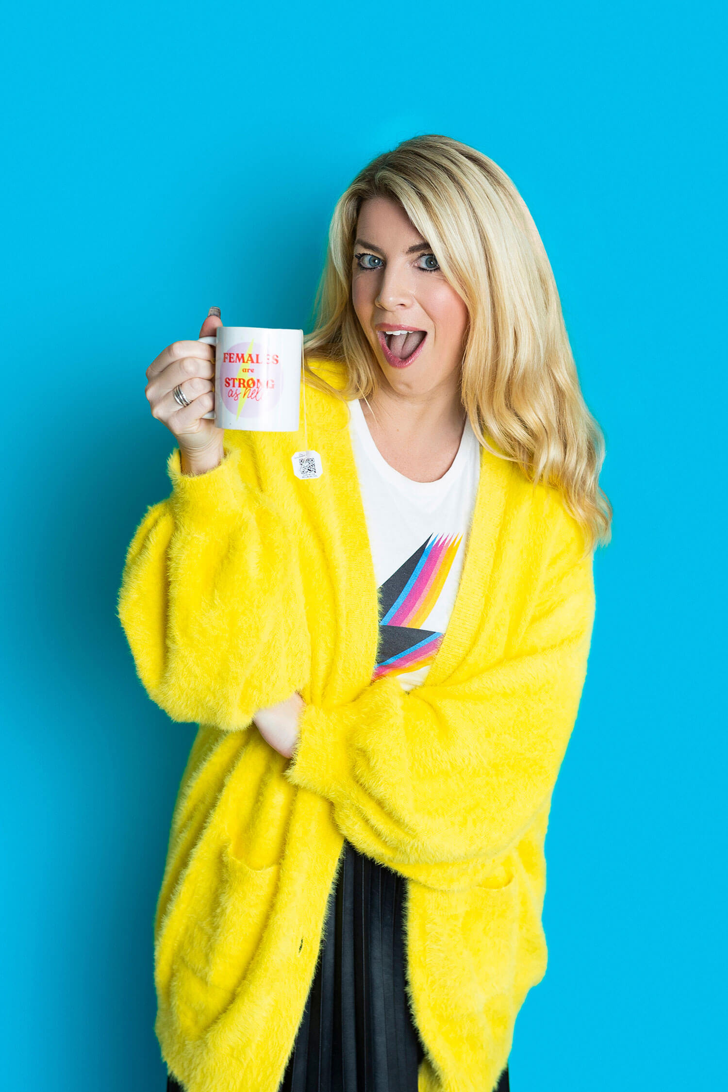 A woman - Laura Brunton - in a bright yellow cardigan holds up a mug with a playful expression, standing against a bold turquoise backdrop during a branding shoot.