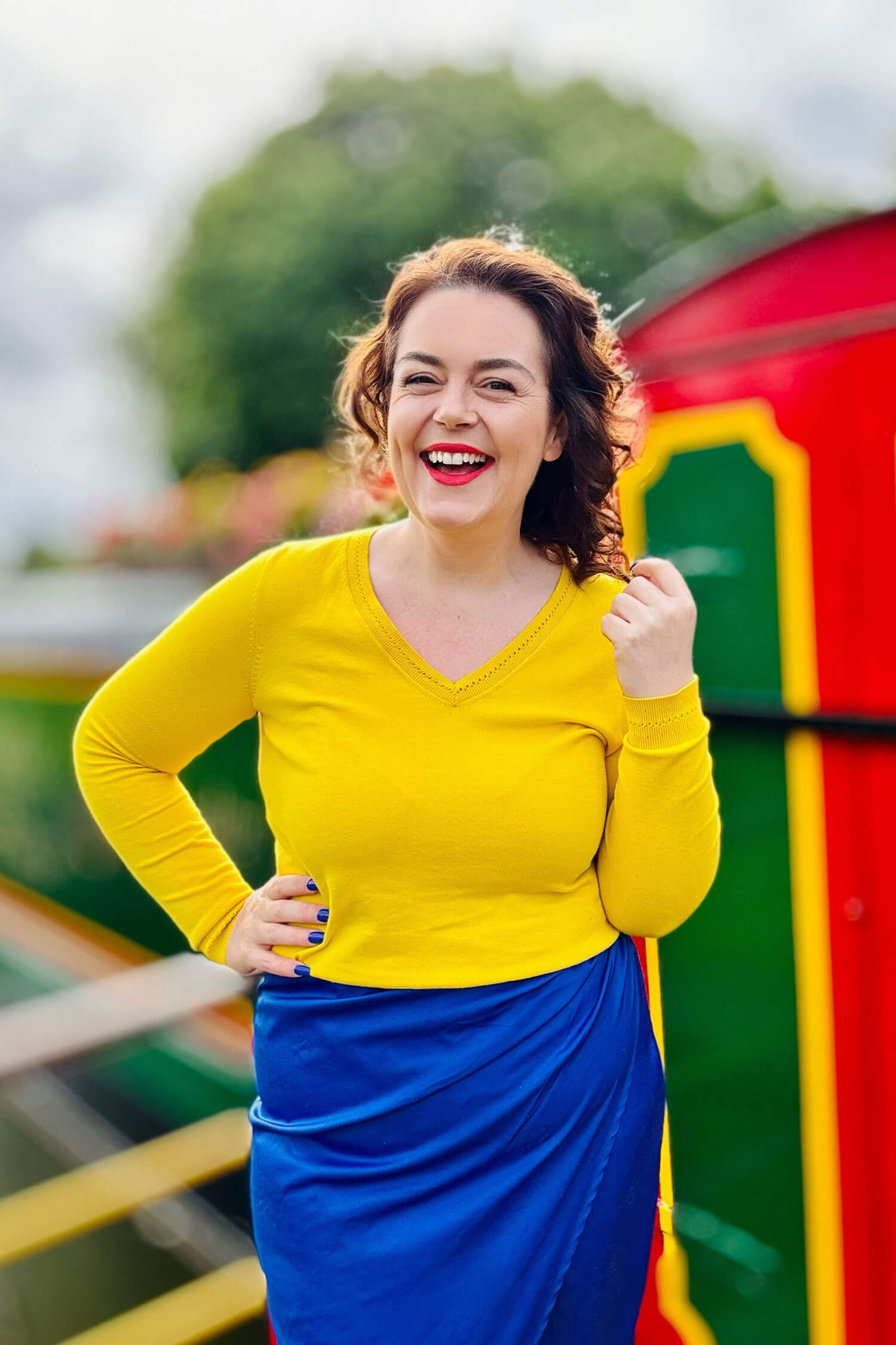 Brand photographer Rosie Parsons smiling brightly outdoors, wearing a yellow top and blue skirt, standing beside a colourful narrowboat with greenery in the background during a business photoshoot taken on her iphone.