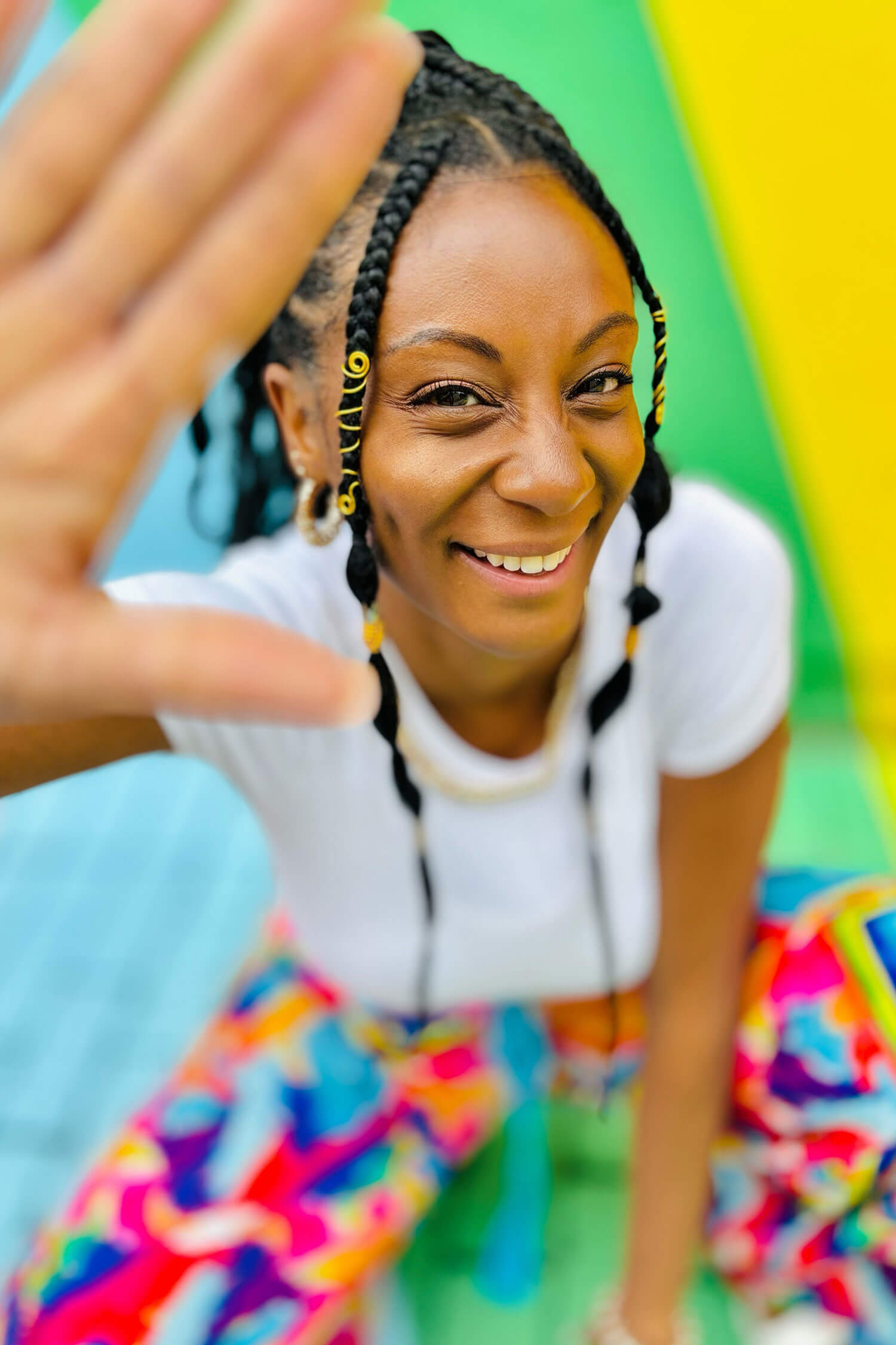 A woman with braided hair poses playfully for the camera, lifting a hand towards the lens while smiling. She’s sitting on a brightly coloured patterned surface, creating a bold, eye-catching look that shows an easy, aesthetic selfie pose for Instagram photos.