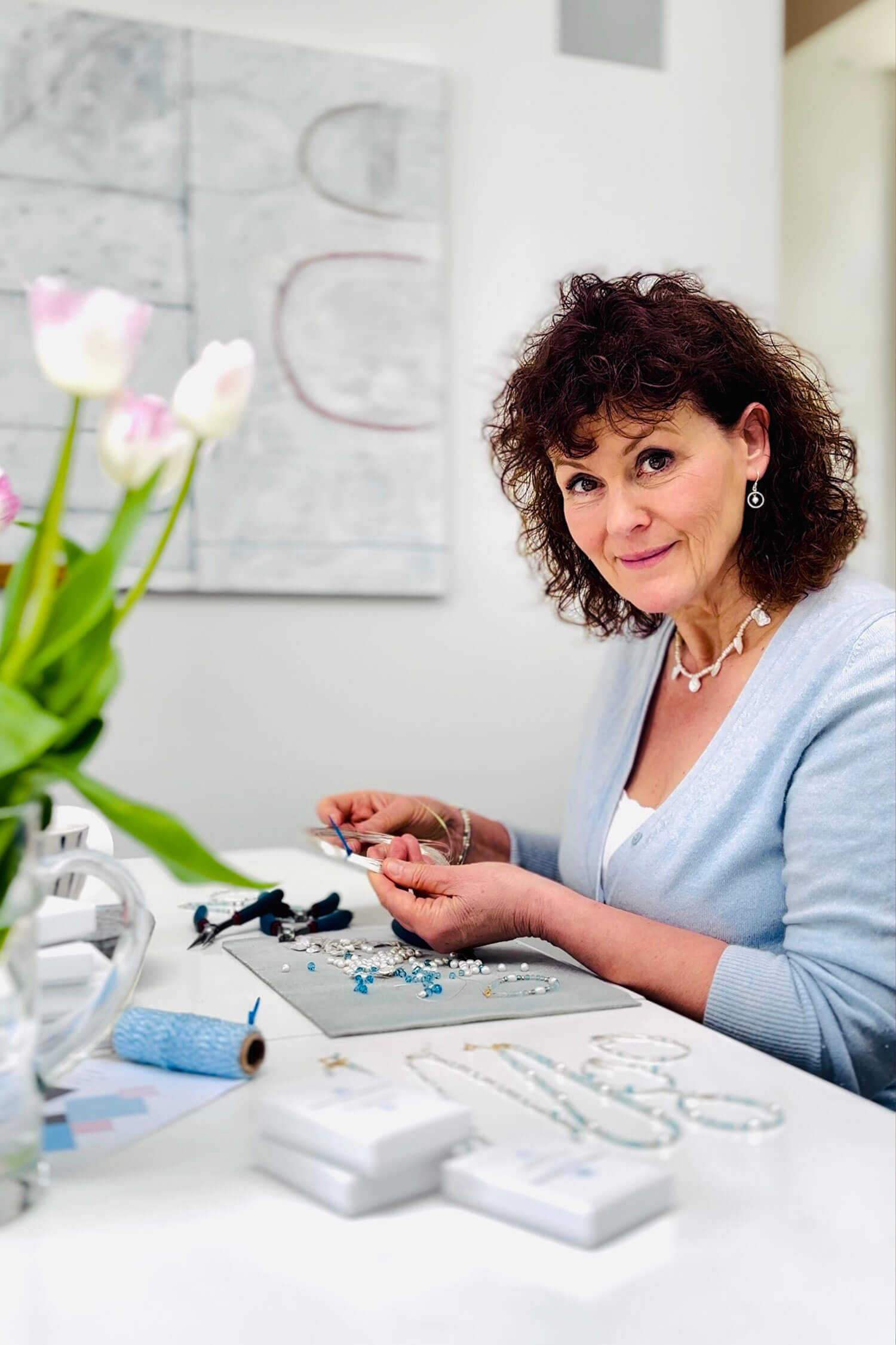 A woman sits at a bright workspace making jewellery by hand, surrounded by beads, tools and finished pieces, looking up with a gentle smile during a self-portrait business photoshoot taken by herself on her own phone.