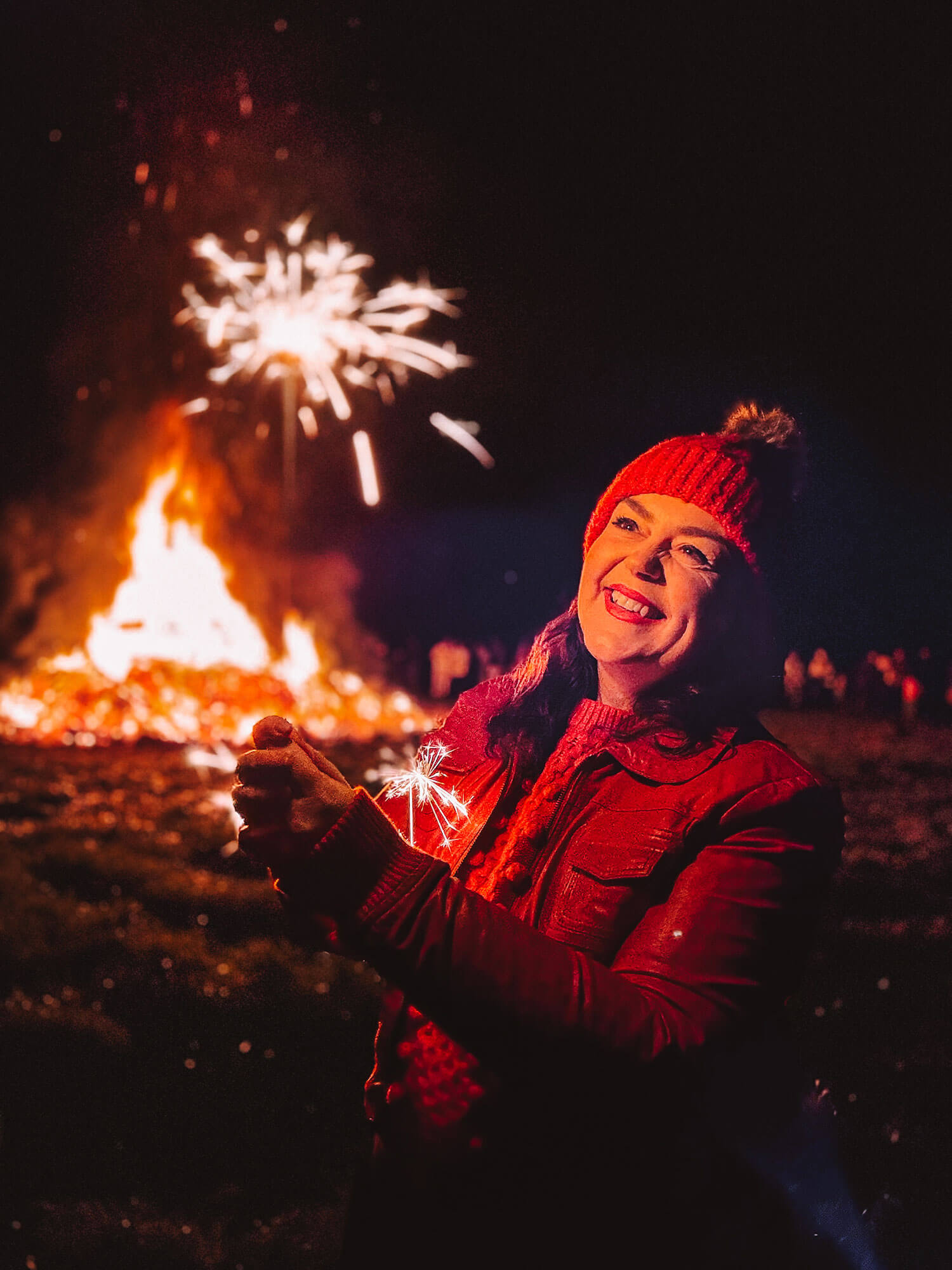 A woman standing outdoors at night smiles while holding a sparkler, with a bonfire and fireworks glowing behind her. The warm autumn colours and festive atmosphere create a cosy seasonal moment that works well for an autumn fall photoshoot