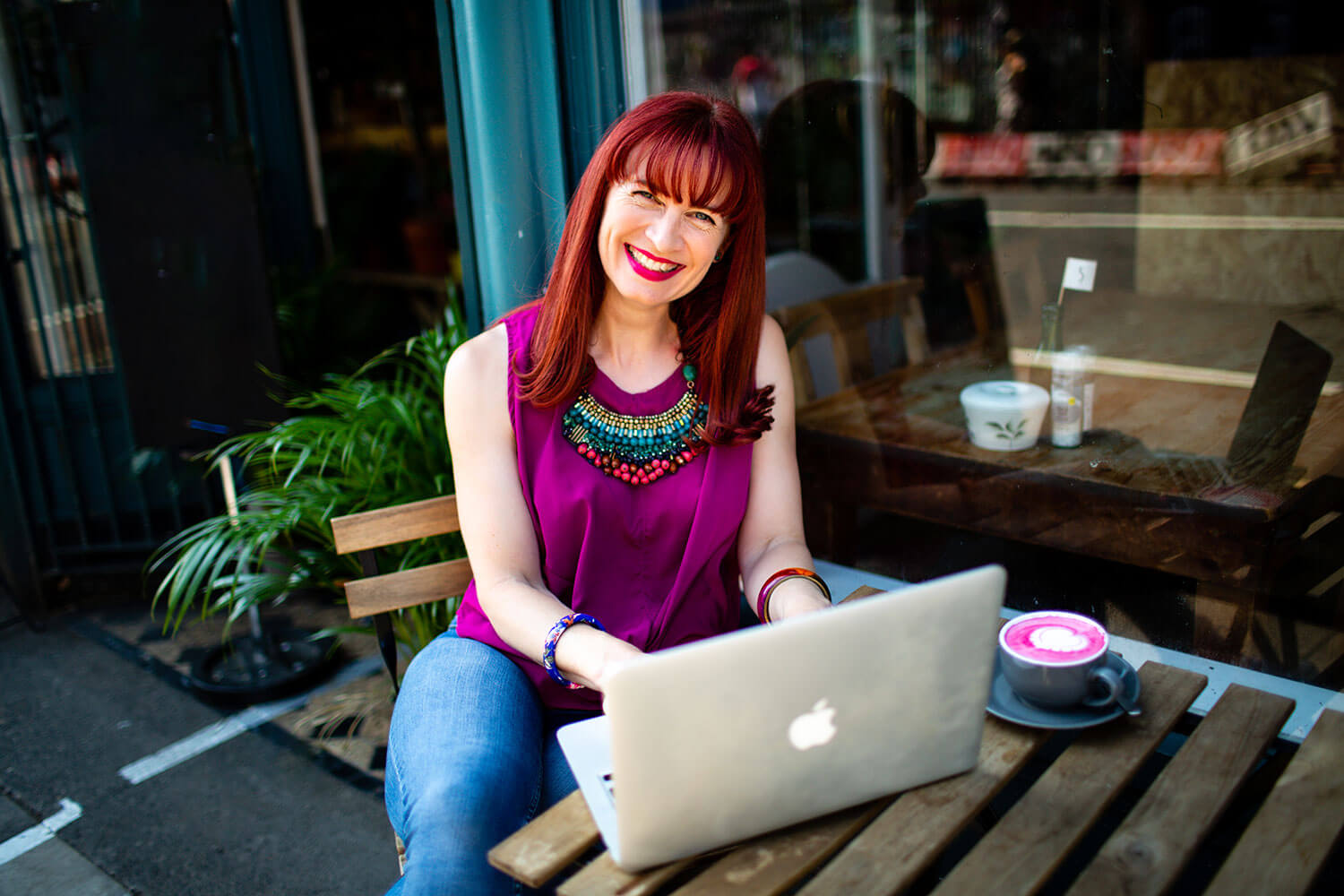 A female entrepreneur with vibrant red hair and a fringe smiles warmly while working on a MacBook laptop at an outdoor cafe. She wears a magenta sleeveless top, blue jeans, colourful bangles, and a striking turquoise and coral statement necklace. A pink beetroot latte in a grey cup sits on the rustic wooden table beside her, with green plants and cafe windows in the background. She's researching the best online course platforms and exploring course website design options for her business.