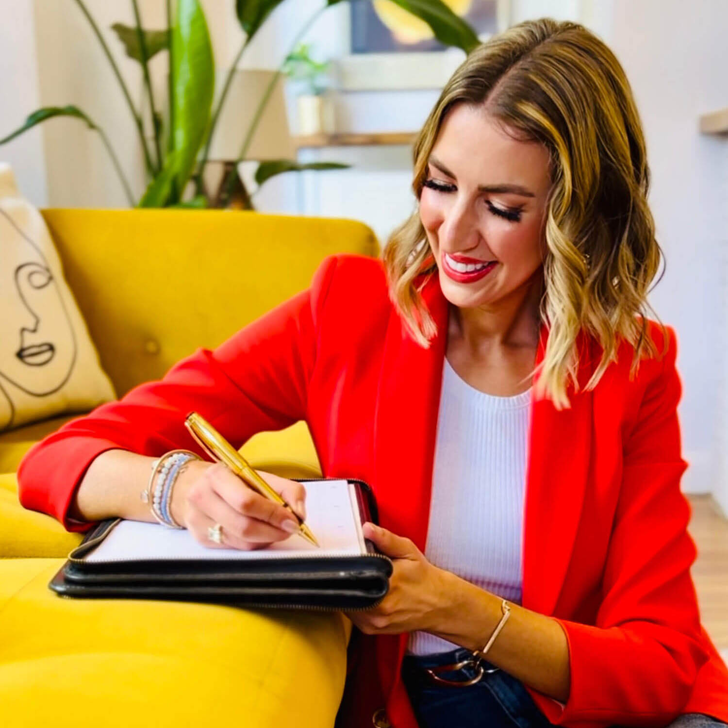 A woman in a red blazer writing in a planner on a yellow sofa with a gold pen showing inspiration for how to start a blog for beginners.