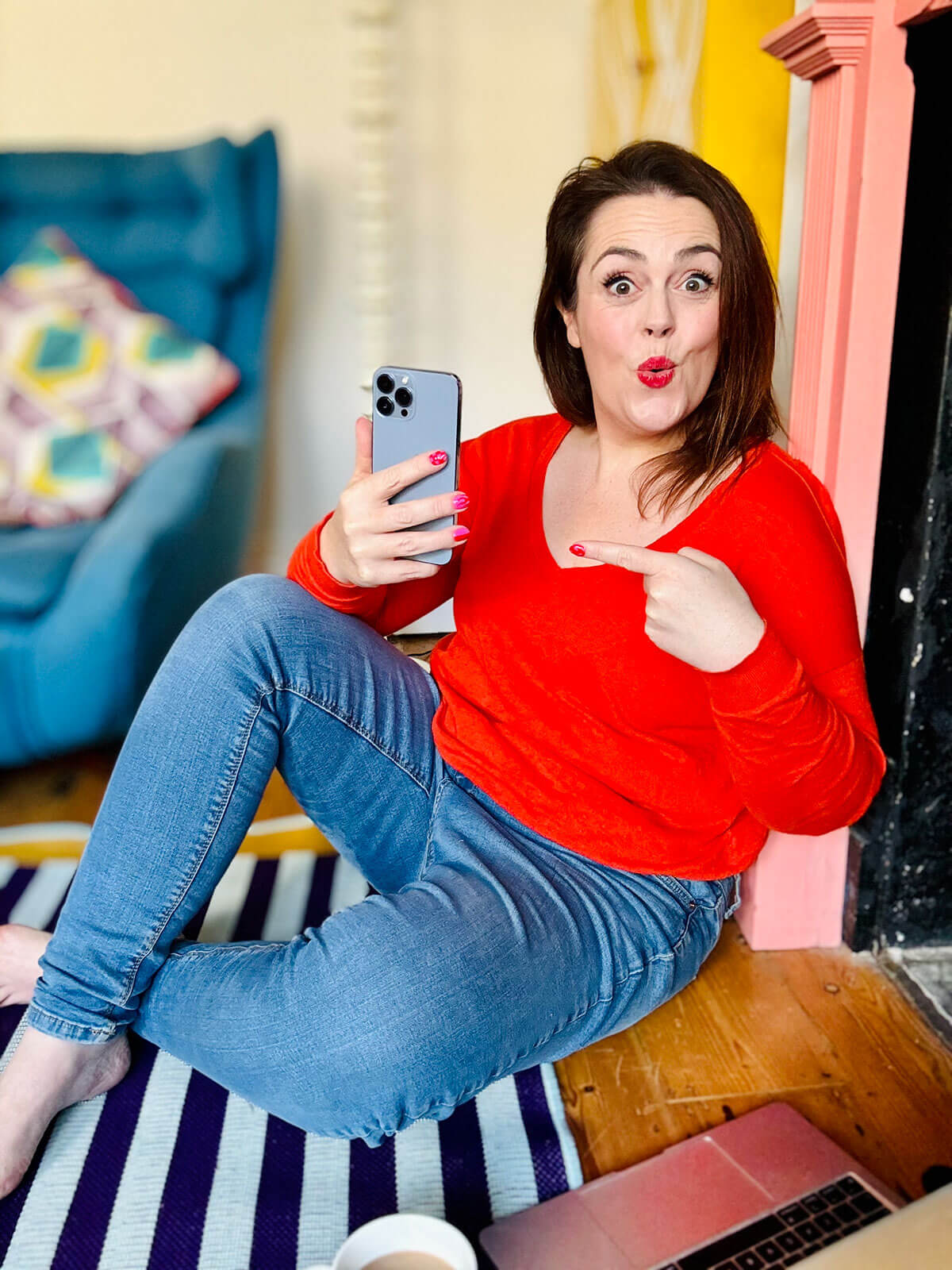Woman in bright red jumper and jeans sitting on floor by a pink fireplace, holding up her phone and pointing at it with a playful expression, representing the best side hustles for women