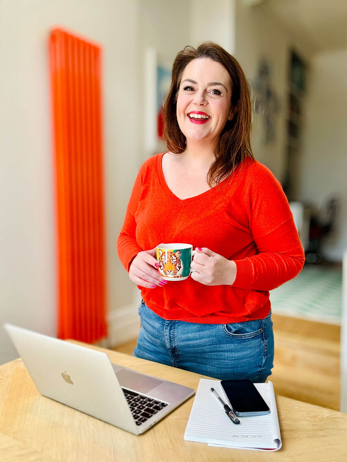 Woman in red v-neck jumper standing at wooden desk holding a tiger print mug, with laptop and notebook ready for work, showing businesses to start from home