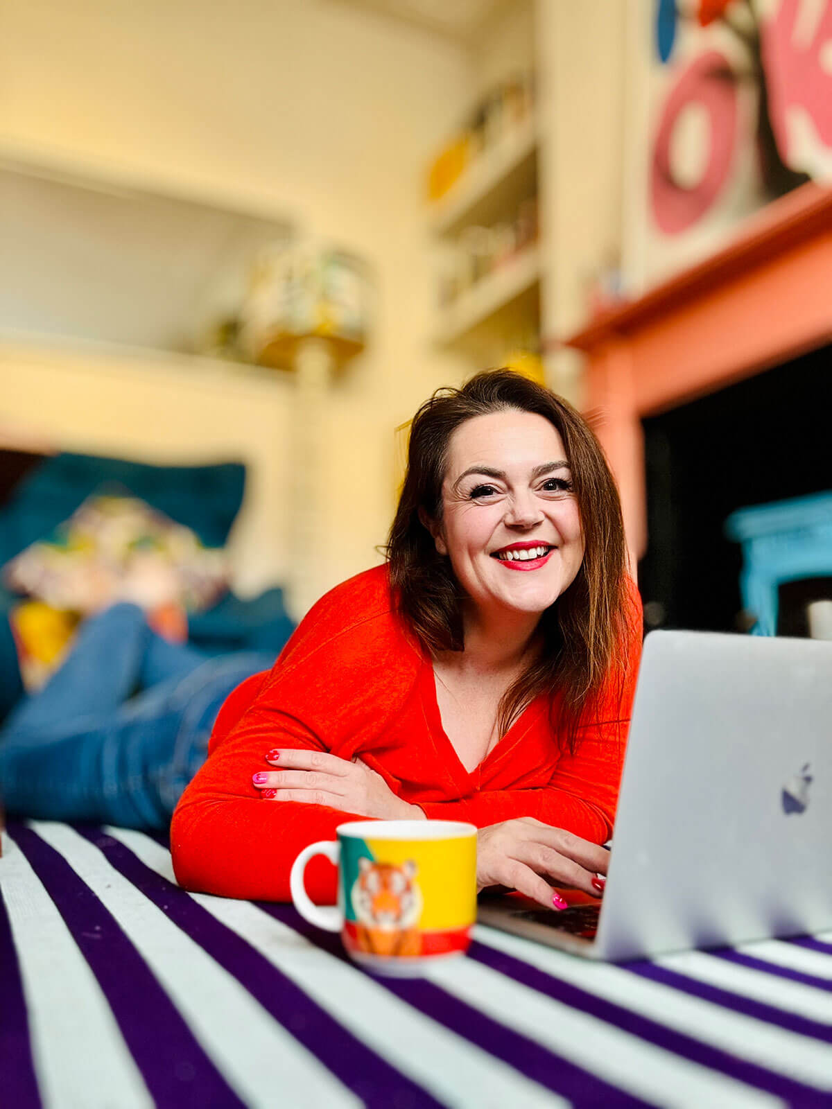 Woman in red top lying on her stomach on a navy striped rug, typing on laptop with tiger mug beside her in cosy living room, demonstrating side hustles from home