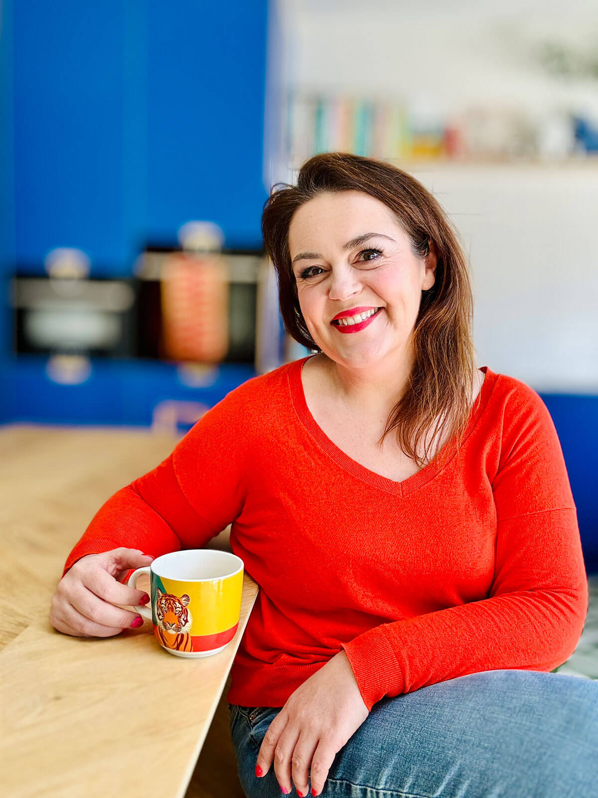 Woman in red jumper sitting at wooden table holding colourful tiger mug, smiling warmly with bright blue kitchen behind her, ready to earn extra money