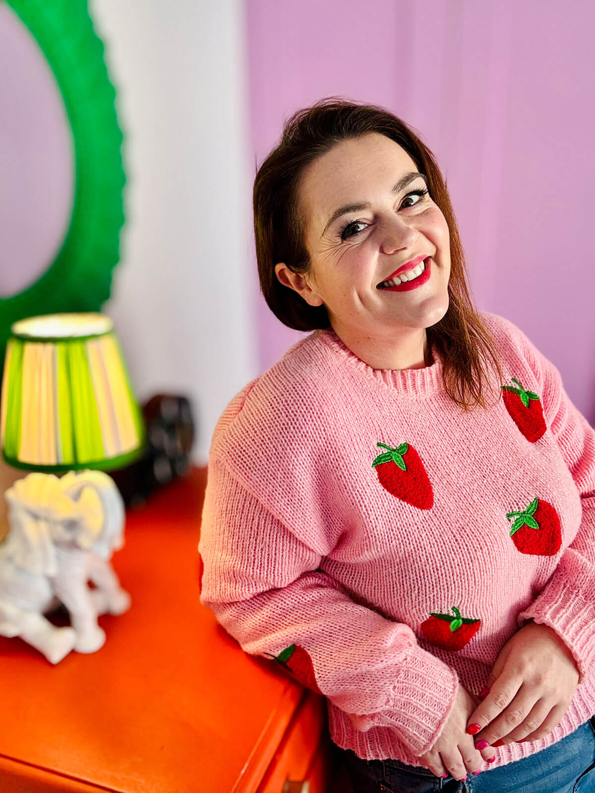 Woman in pink jumper with strawberry pattern sitting on orange dresser, smiling and looking to the side in colourful room with green mirror and striped lamp, building extra income