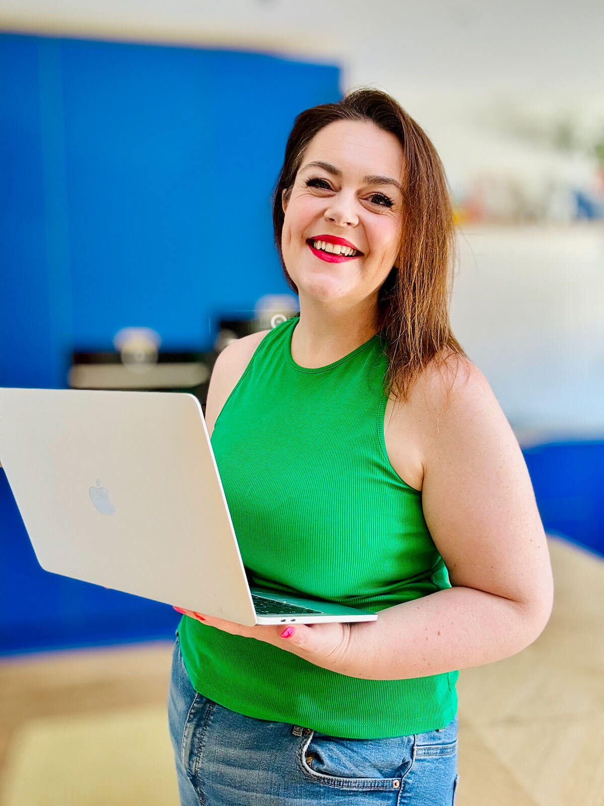 Woman in green sleeveless top standing and holding open laptop while smiling broadly, blue kitchen blurred in background, enjoying easy online jobs