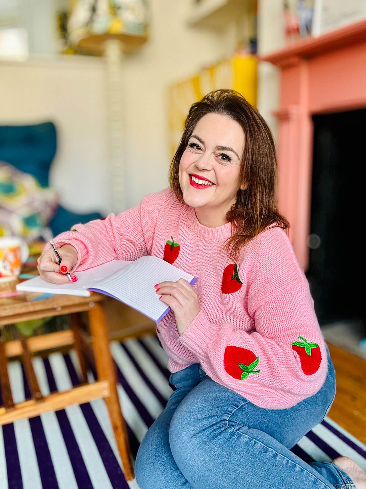 Woman in pink strawberry jumper kneeling on striped rug writing in notebook with pen, living room with pink fireplace behind, representing jobs for women