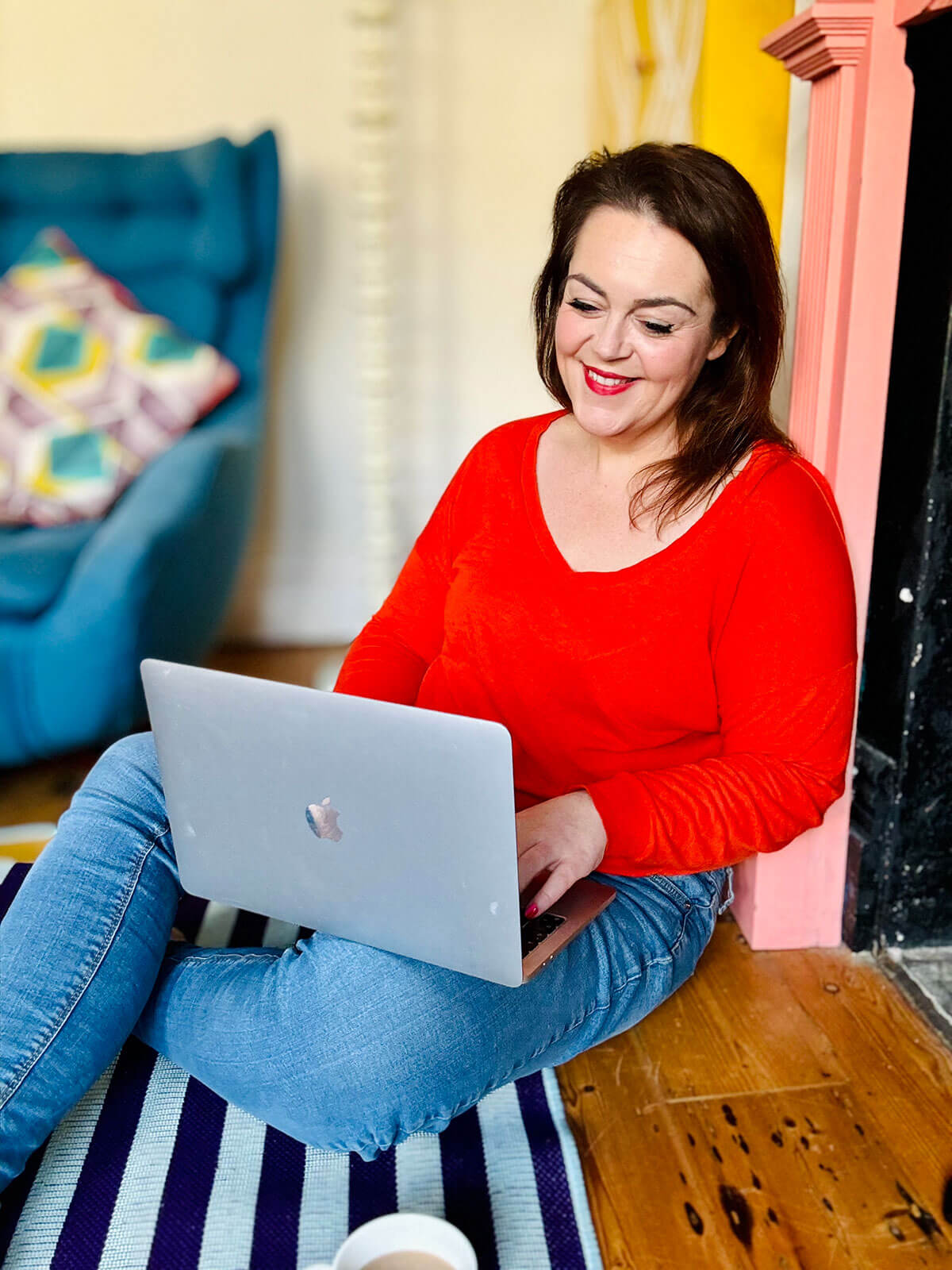 Woman in red top sitting cross-legged on wooden floor by pink fireplace, laptop balanced on lap, focused on screen with teal chair in background, working on side hustle business ideas