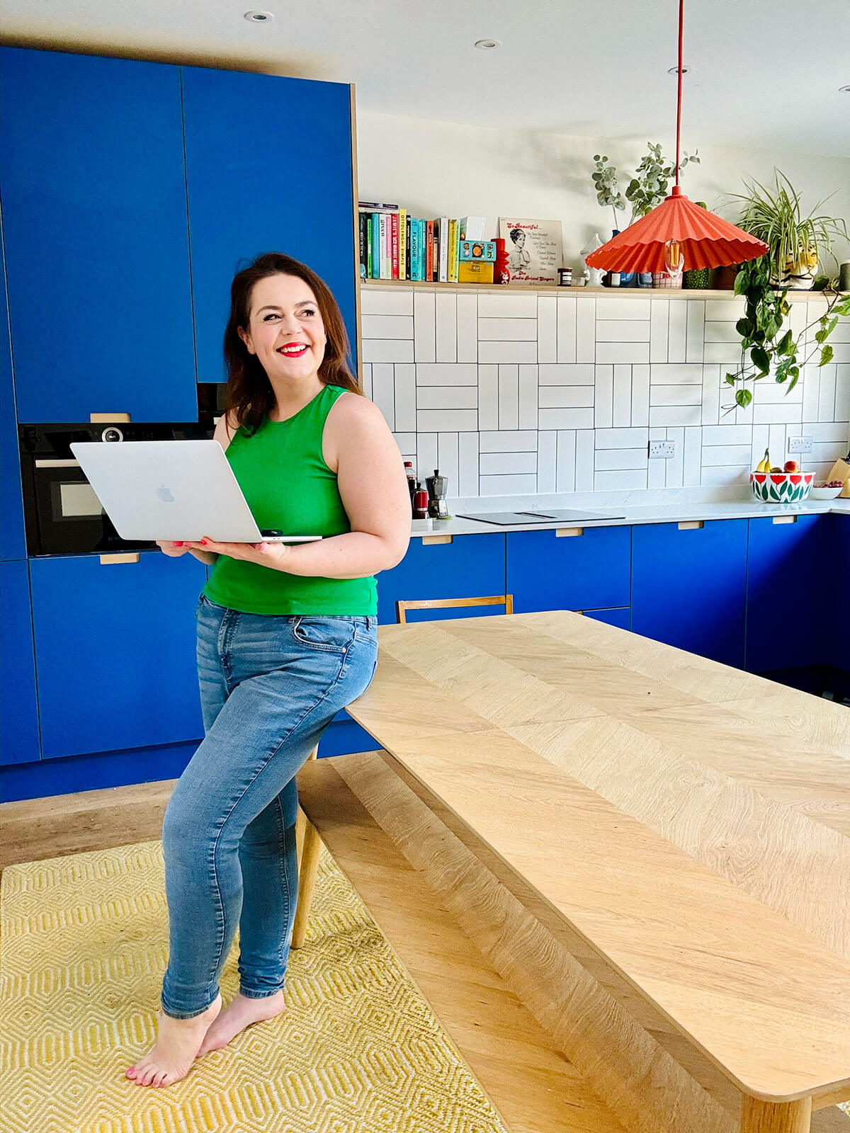 Woman in green top standing barefoot in bright blue kitchen holding laptop, large wooden table in foreground, plants and cookbooks on shelves, researching side hustle ideas