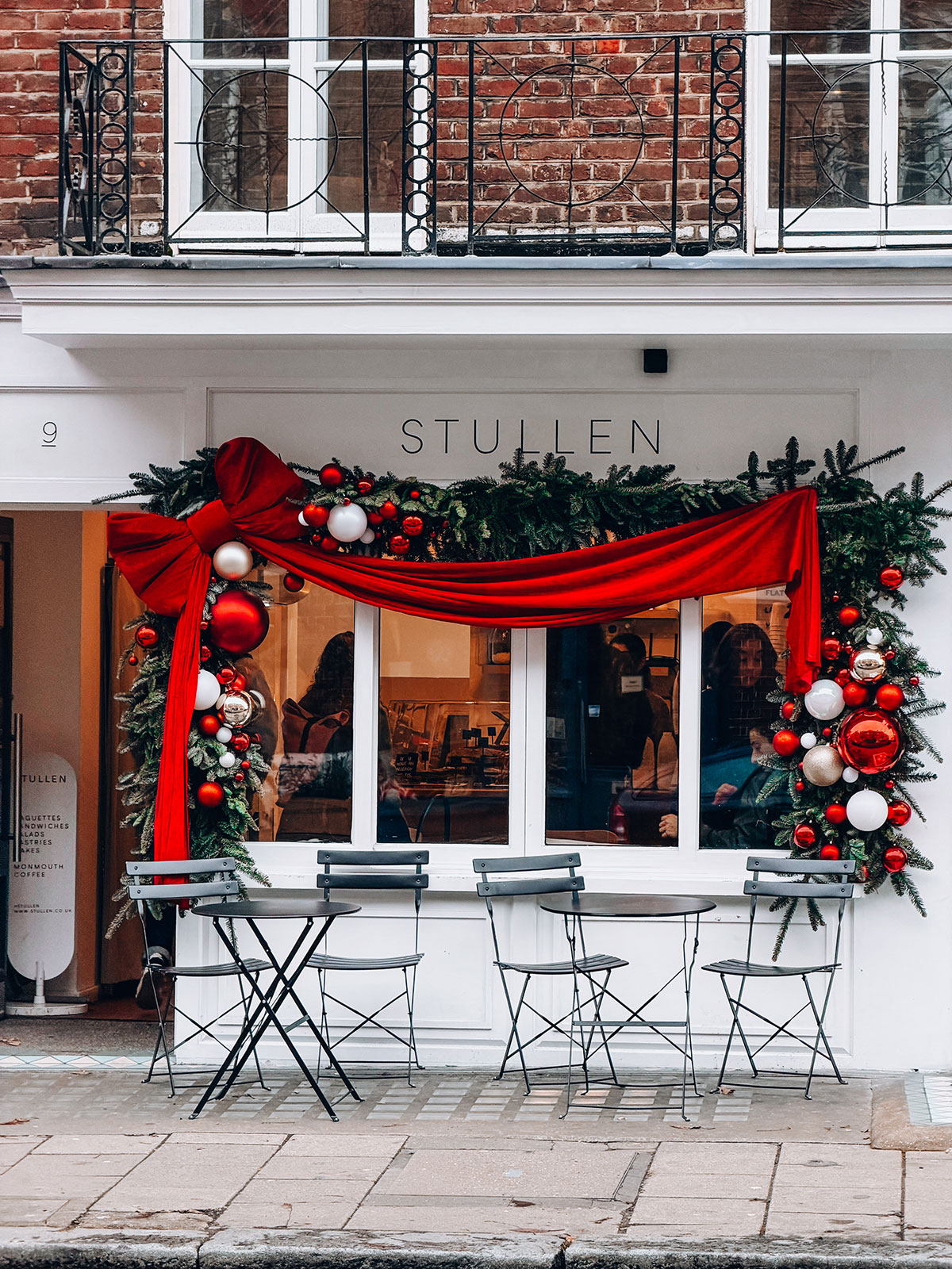 A white cafe front decorated for Christmas with pine garlands, red and white baubles and a large red ribbon bow draped across the window. Grey metal bistro tables and chairs sit outside on the pavement. The scene shows a vintage christmas photoshoot location with beautiful festive styling.