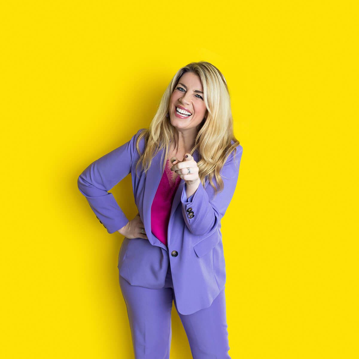 Laura Brunton business coach laughing while pointing at the camera, hand on hip, purple suit against yellow background
