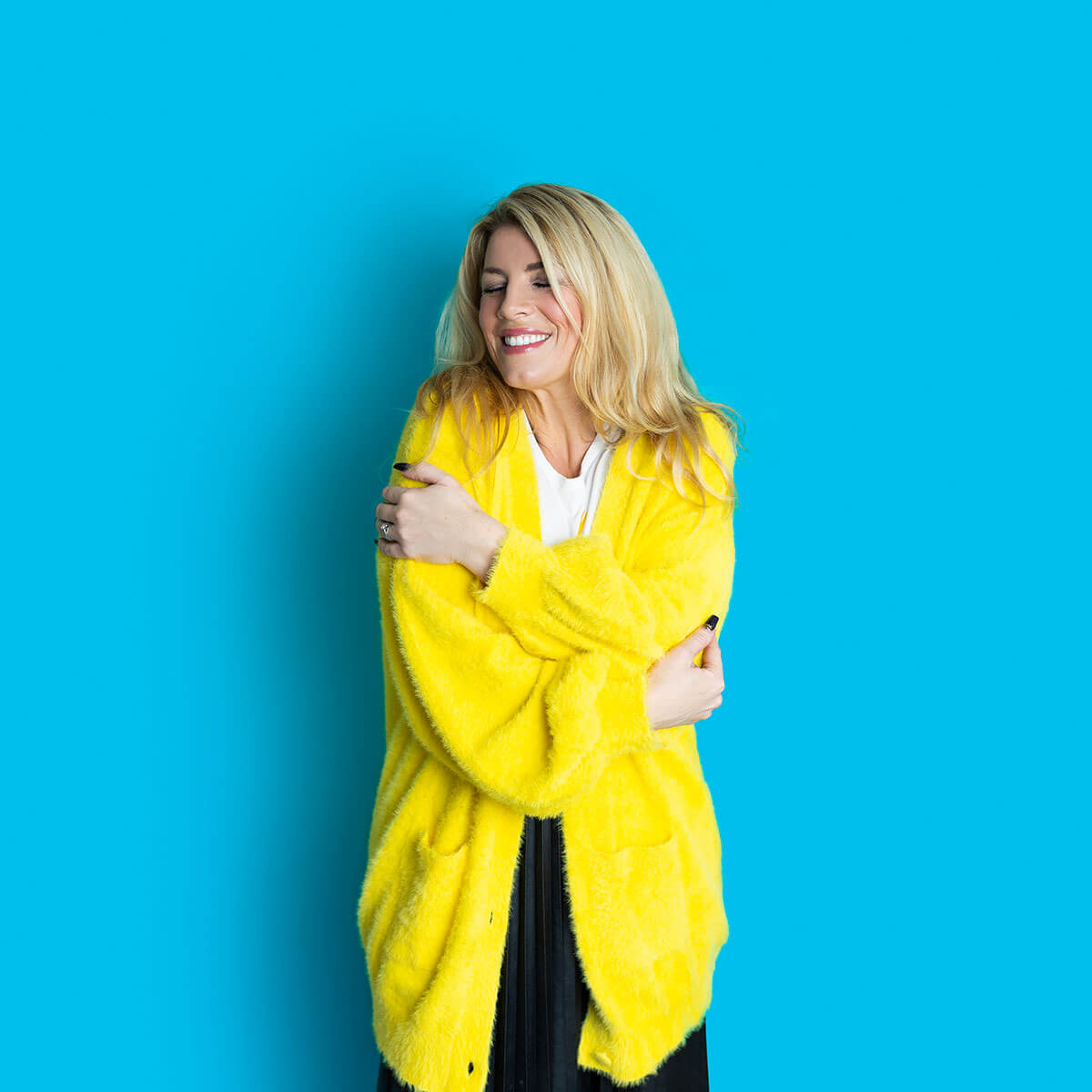 Laura Brunton business coach smiling with eyes closed and hugging herself in a yellow coat against a blue background during a studio branding photoshoot