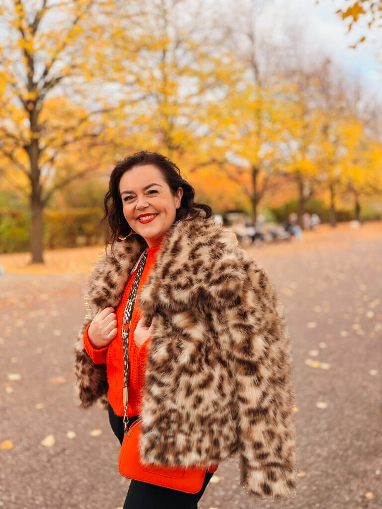 Rosie Parsons, brand photographer and business blogger, smiling on an autumn walk in a park with golden yellow trees in the background. She wears a leopard print faux fur coat over a bright orange jumper with an orange crossbody bag. Fallen leaves scatter the path around her. An image representing self care routine ideas, healthy lifestyle habits and getting outdoors for your mental and emotional health.