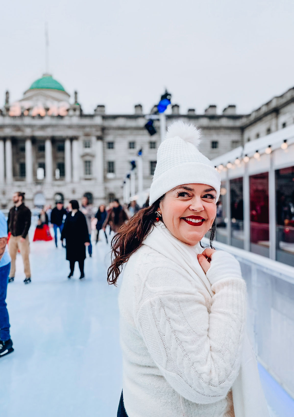 Rosie Parsons, headshot photographer and business blogger, smiling while ice skating at Somerset House in London. She wears an all-white outfit with a cream cable-knit sweater, white bobble hat and white scarf. The historic building is visible behind her, demonstrating christmas fashion photography with winter styling.