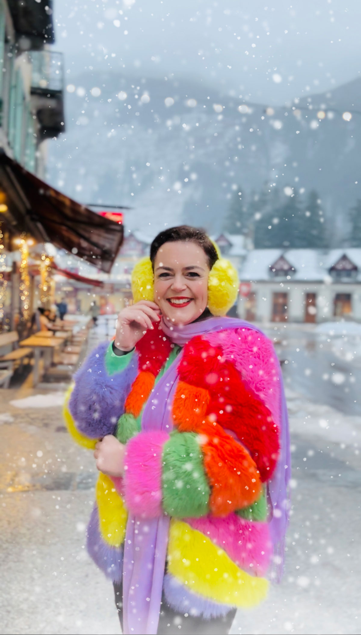 Rosie Parsons, professional photographer and business blogger, smiling in falling snow in a snowy mountain village. She wears a colourful rainbow faux fur coat, lilac scarf and bright yellow ear muffs. Snow-covered chalets and twinkling lights are visible behind her, showing a creative christmas photoshoot in a winter setting.