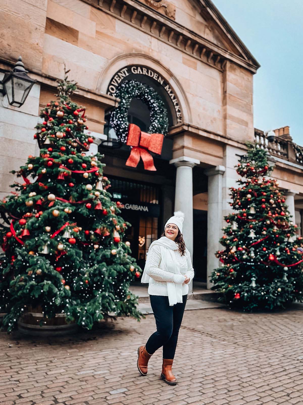 Rosie Parsons, brand photographer and business blogger, smiling in front of Covent Garden Market in London. She wears a cream cable-knit sweater, white bobble hat, white scarf and tan boots. Two large decorated Christmas trees with red and gold baubles and a giant wreath with red bow frame the entrance, showing a vintage christmas photoshoot spot.