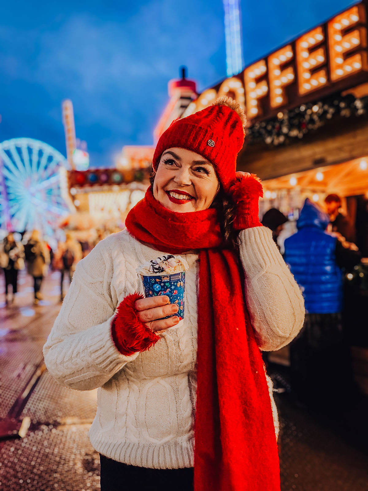 Rosie Parsons, professional photographer and business blogger, smiling at a Christmas market while holding a hot chocolate with whipped cream. She wears a white cable-knit sweater, red beanie, red scarf and red gloves. A lit-up coffee sign glows behind her with a ferris wheel in the background, showing a cozy christmas photoshoot setup at dusk.