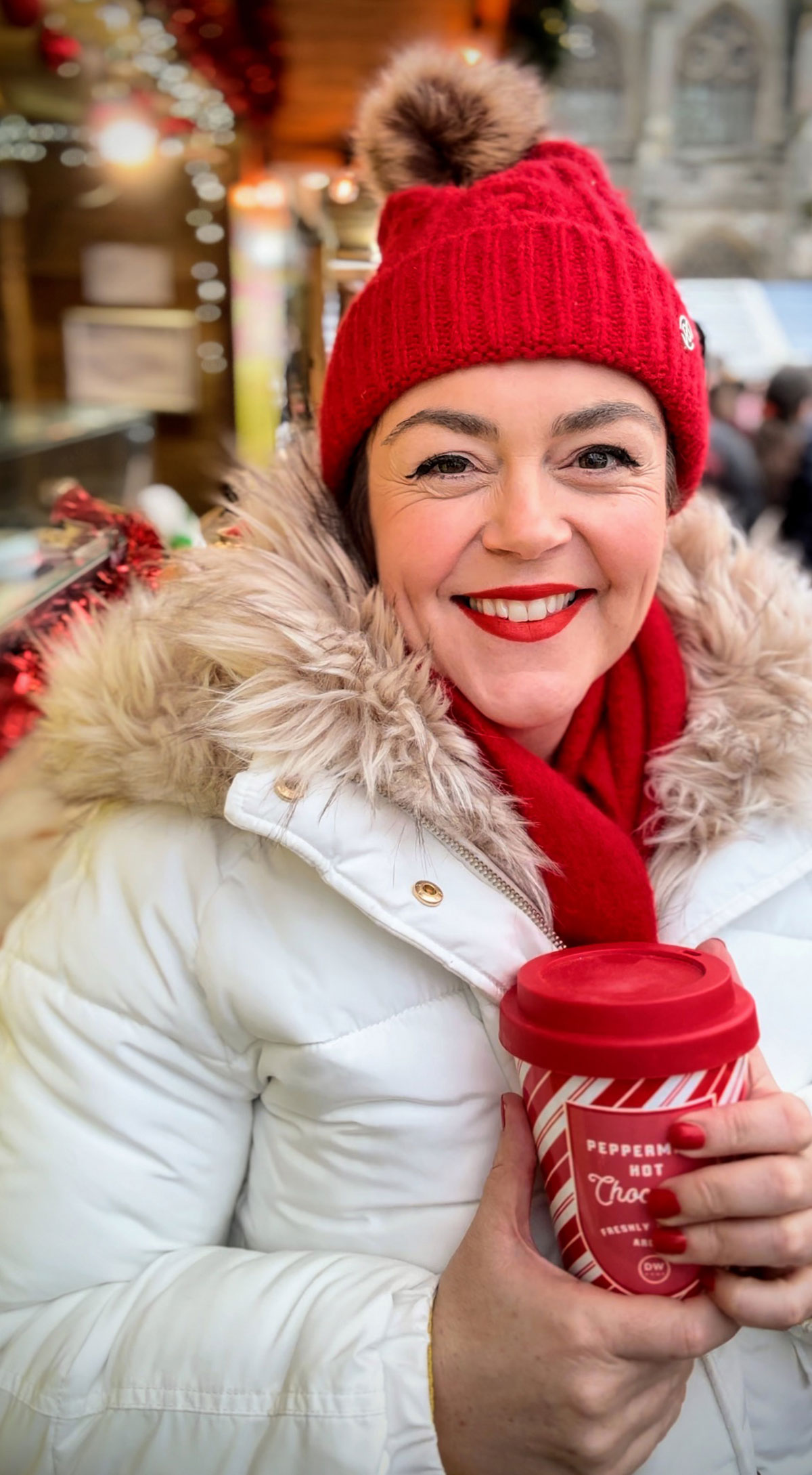 Rosie Parsons, headshot photographer and business blogger, smiling while holding a peppermint hot chocolate at a Christmas market. She wears a white puffer coat with faux fur hood, red bobble hat and red scarf. Twinkly lights and festive decorations glow behind her, demonstrating christmas poses for a festive photoshoot.