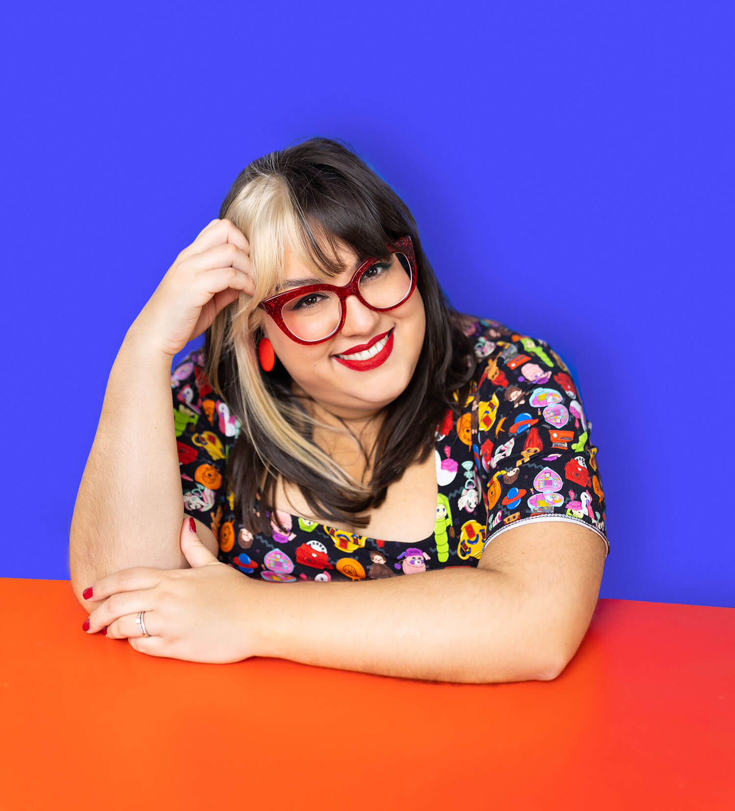 Smiling woman with two-toned hair and bold red glasses leans on a bright red table against a vivid blue background. Her statement frames and expressive style highlight how glasses can enhance recognisability and reflect personality in a professional headshot.