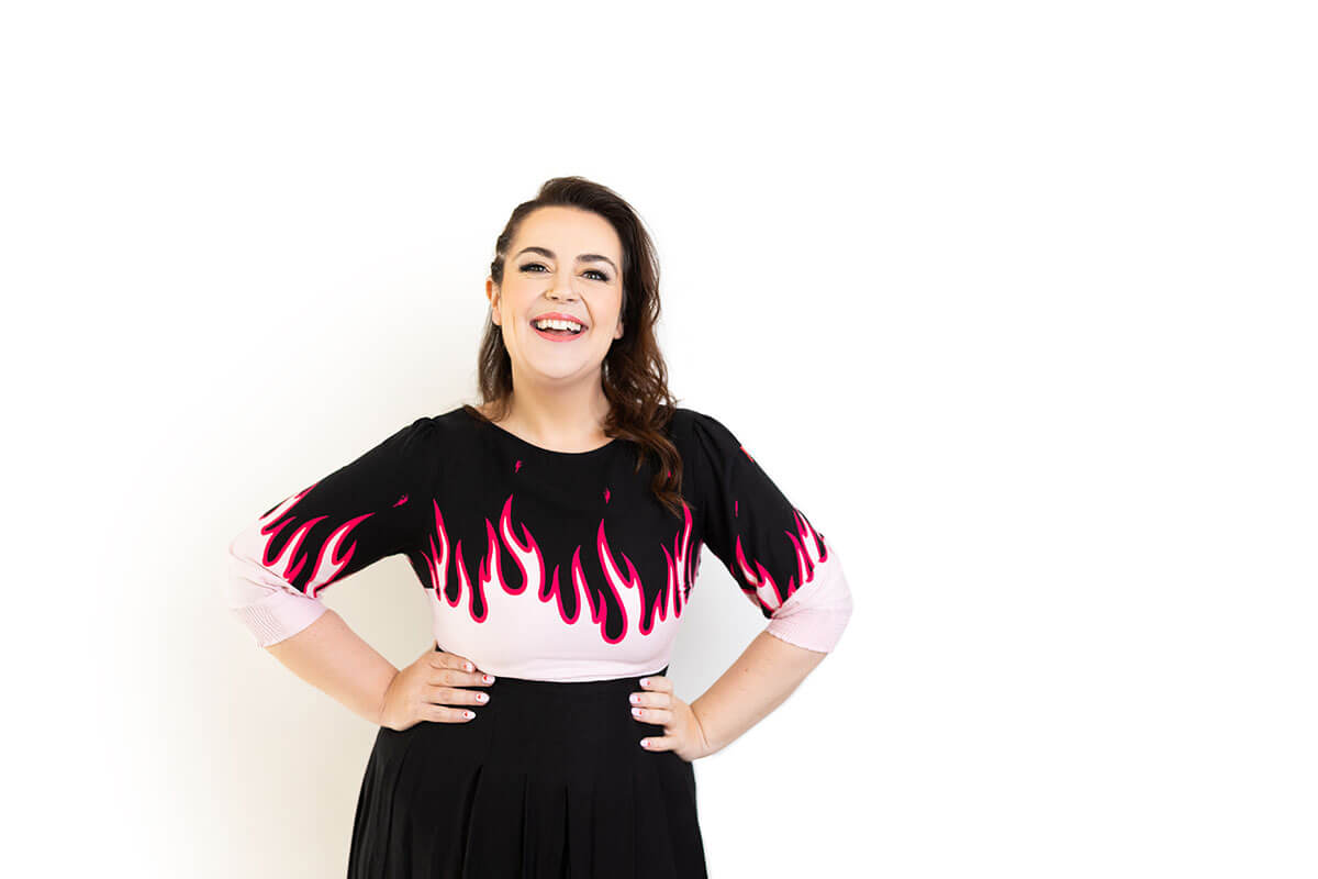 Confident woman laughing in a black and pink top with flame graphics, paired with a pleated black skirt, standing against a clean white background. Her bold outfit and expressive pose reflect a vibrant brand personality, perfect for a company website headshot that aligns with personal branding.