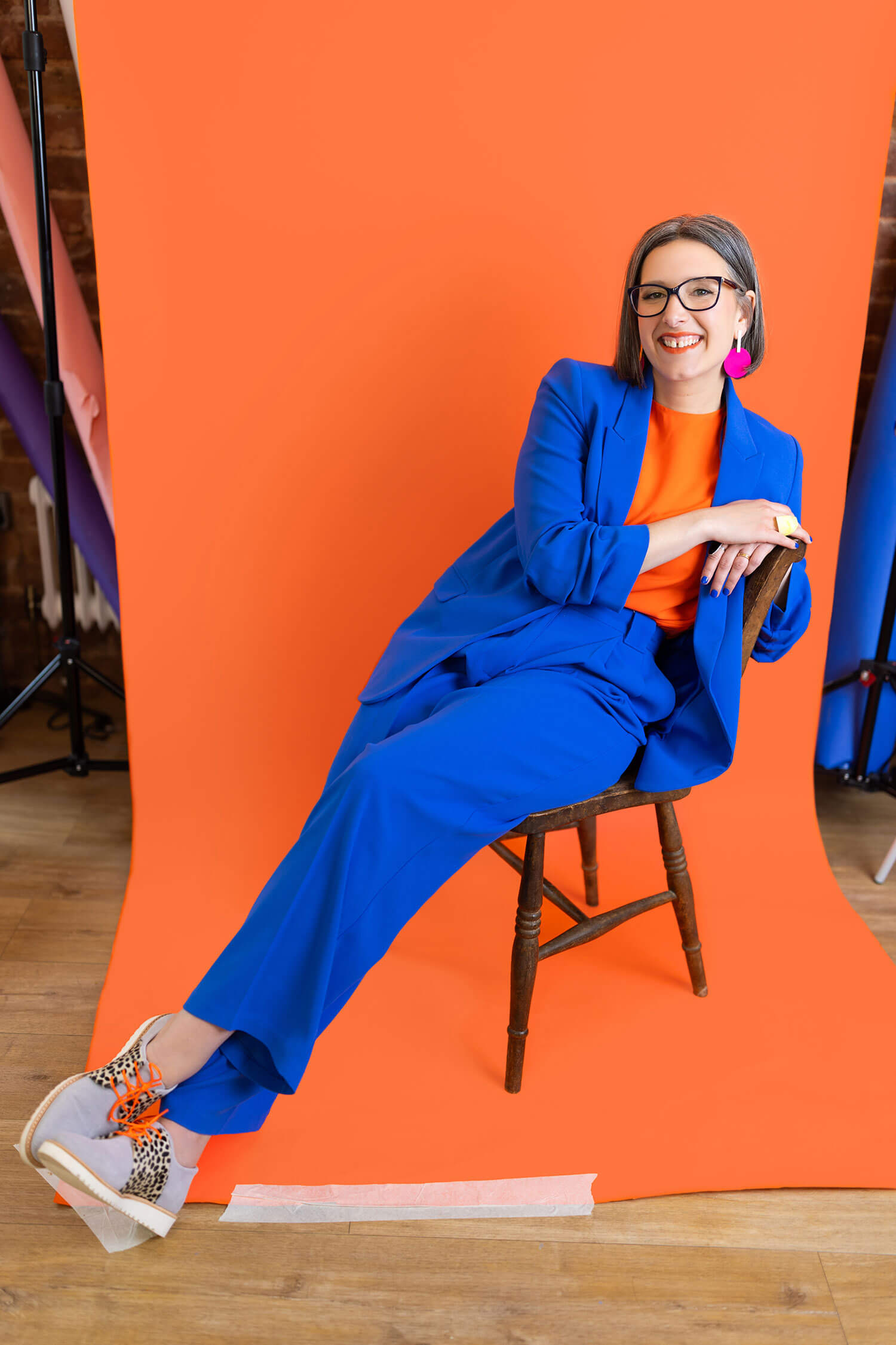 Smiling woman in a vibrant cobalt blue suit and orange top leans confidently on a wooden chair against an orange studio backdrop. Her bold, tailored outfit and relaxed pose strike the perfect balance of authority and personality for speaker bios or media features.