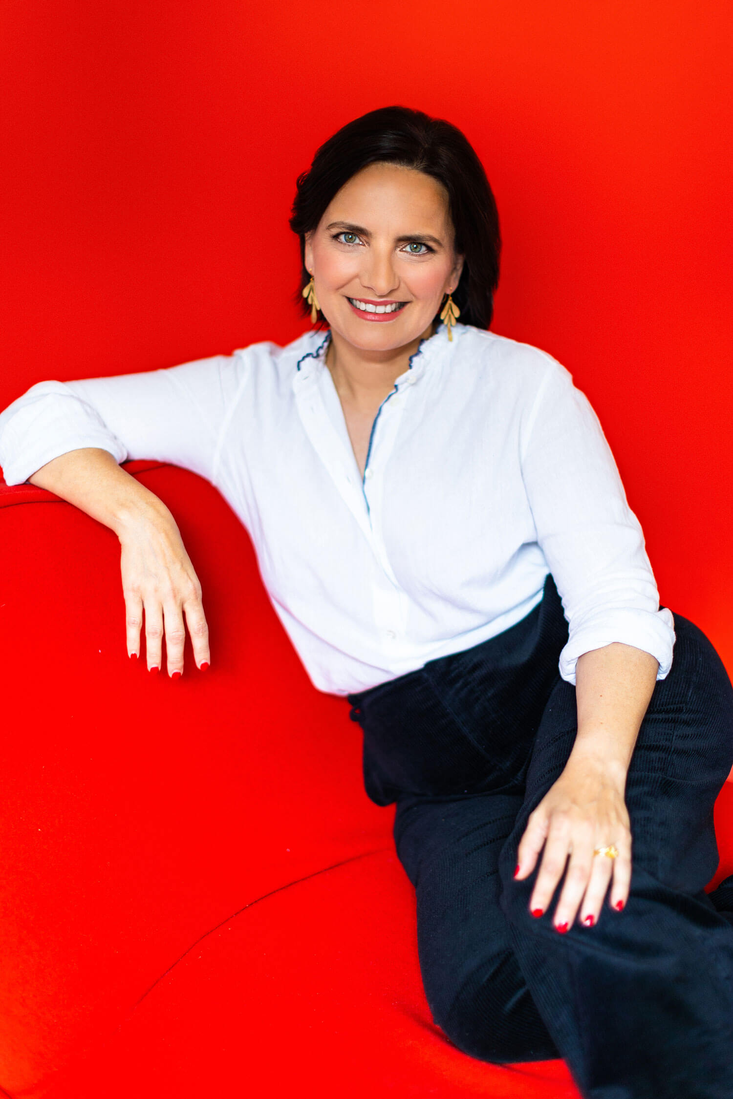 Smiling lawyer in a white blouse and black trousers sits on a vibrant red sofa, combining warmth and professionalism against a bold red background. Her outfit reflects the approachable yet trustworthy look clients expect from a legal professional in a headshot.
