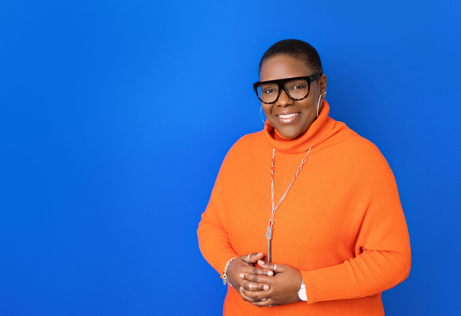 Smiling woman wearing an orange turtleneck sweater and bold black glasses stands in front of a vivid blue background. Her outfit draws attention to her face and neckline, illustrating how headshots focus on the upper frame and why clothing near the face matters most.