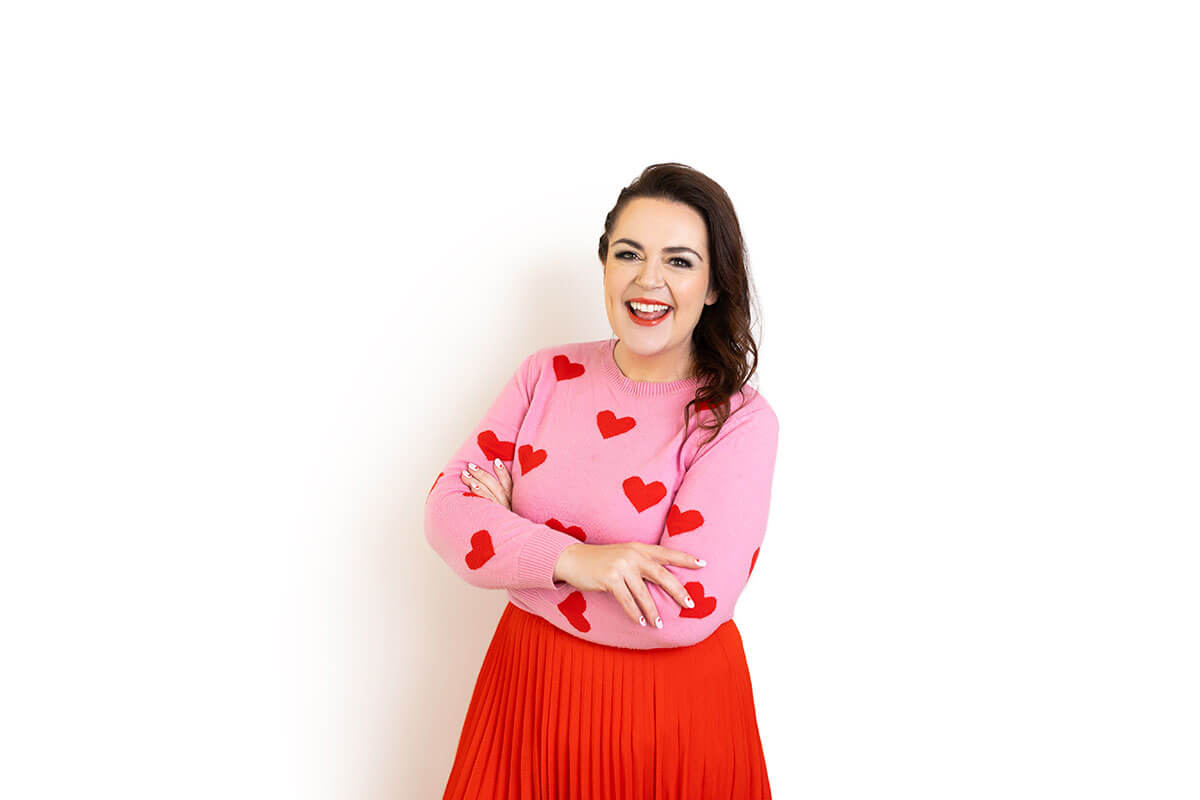 Confident woman in a pink sweater with red hearts and a bright red pleated skirt smiles warmly with arms crossed against a white background. Her bold, fitted outfit and joyful expression reflect how posture, colour, and comfort help you look like the most confident version of yourself in a headshot.