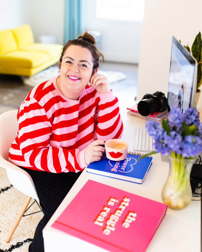 Brand photographer Rosie Parsons at her desk wearing a pink and red striped top, holding a coffee cup beside an iMac, camera, and purple flowers.
