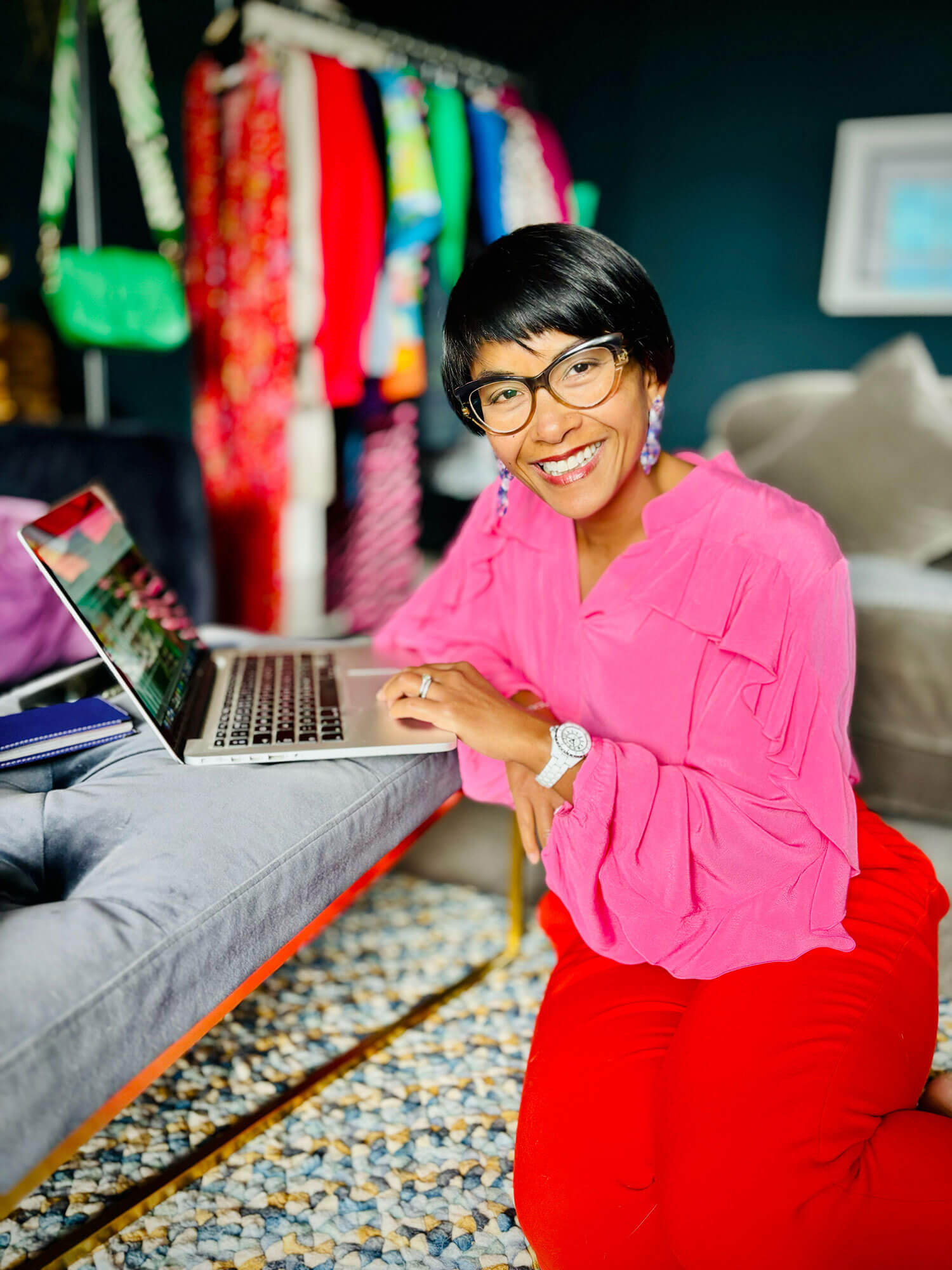 Personal stylist Chantelle Znideric in a pink blouse and red trousers smiles while working on her blog on her laptop, with a colourful clothing rail in the background 