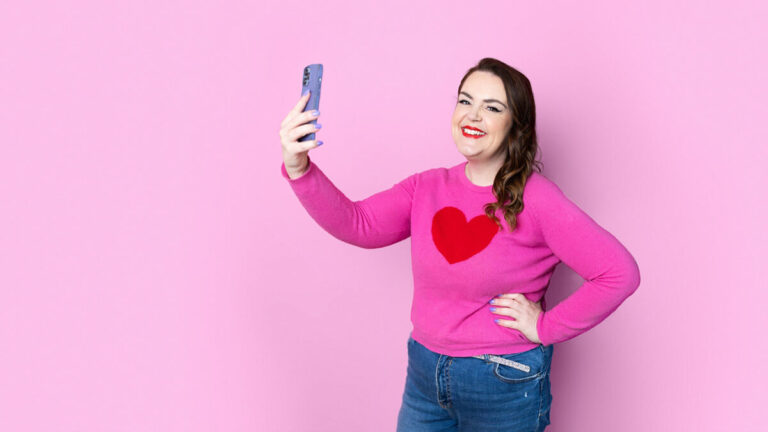 Woman confidently taking a selfie with smartphone held high, hand on hip, wearing pink sweater with red heart and jeans against pink background, demonstrating proper phone angle and body positioning for flattering self portraits