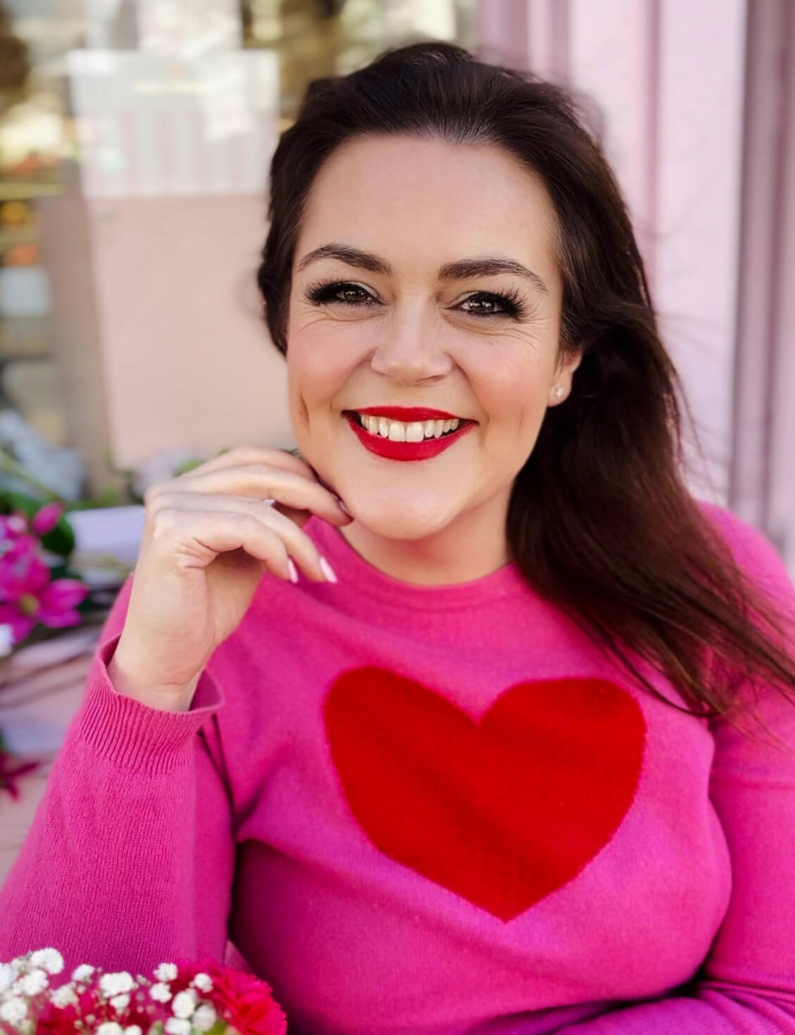 Smiling woman with dark hair poses in natural light wearing a pink sweater with red heart, hand gently resting near her chin, demonstrating a relaxed selfie pose with soft pink background and flowers