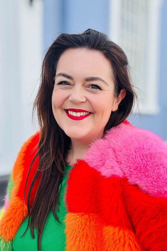 Woman (Rosie P) with dark hair smiling wearing colourful rainbow faux fur coat, photographed against simple clean blue building backdrop – demonstrating how an uncluttered background makes the subject stand out in self portraits in Rosie's guide on how to take a good selfie