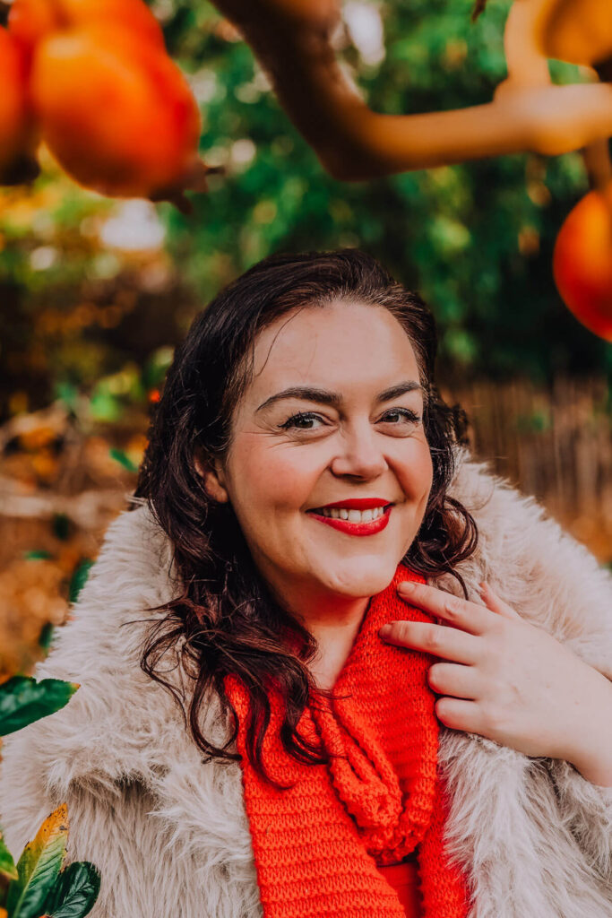 Woman (Rosie Parsons) with dark curly hair smiling outdoors in autumn setting, hand gently touching orange knitted scarf, wearing cream faux fur coat with persimmon fruit tree in background – demonstrating how adding hand movement creates natural-looking self portraits