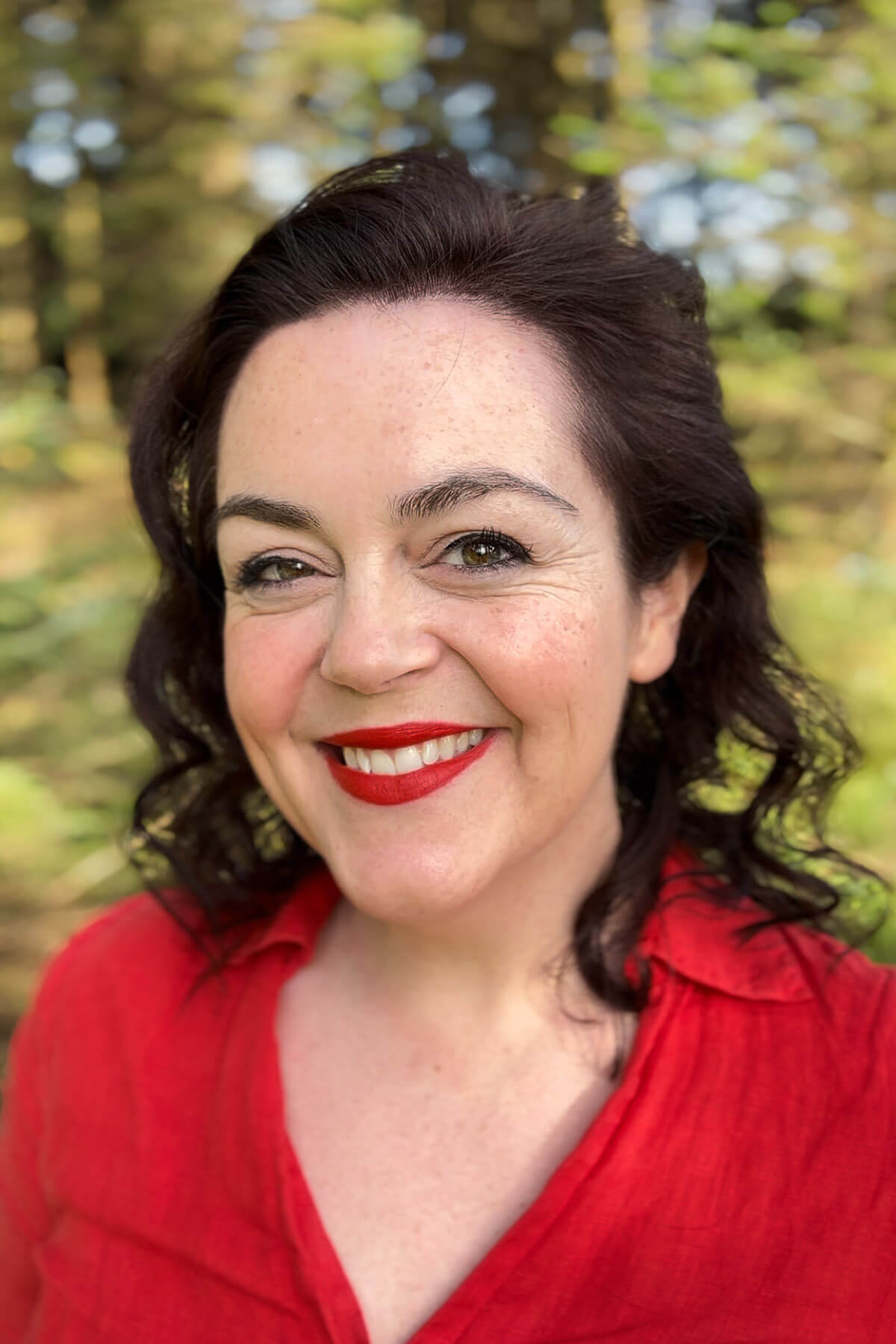 Woman (Rosie Parsons) with dark curly hair smiling warmly wearing red blouse, photographed outdoors with beautifully blurred green bokeh forest background – demonstrating professional-looking self portrait with flattering natural light and genuine expression